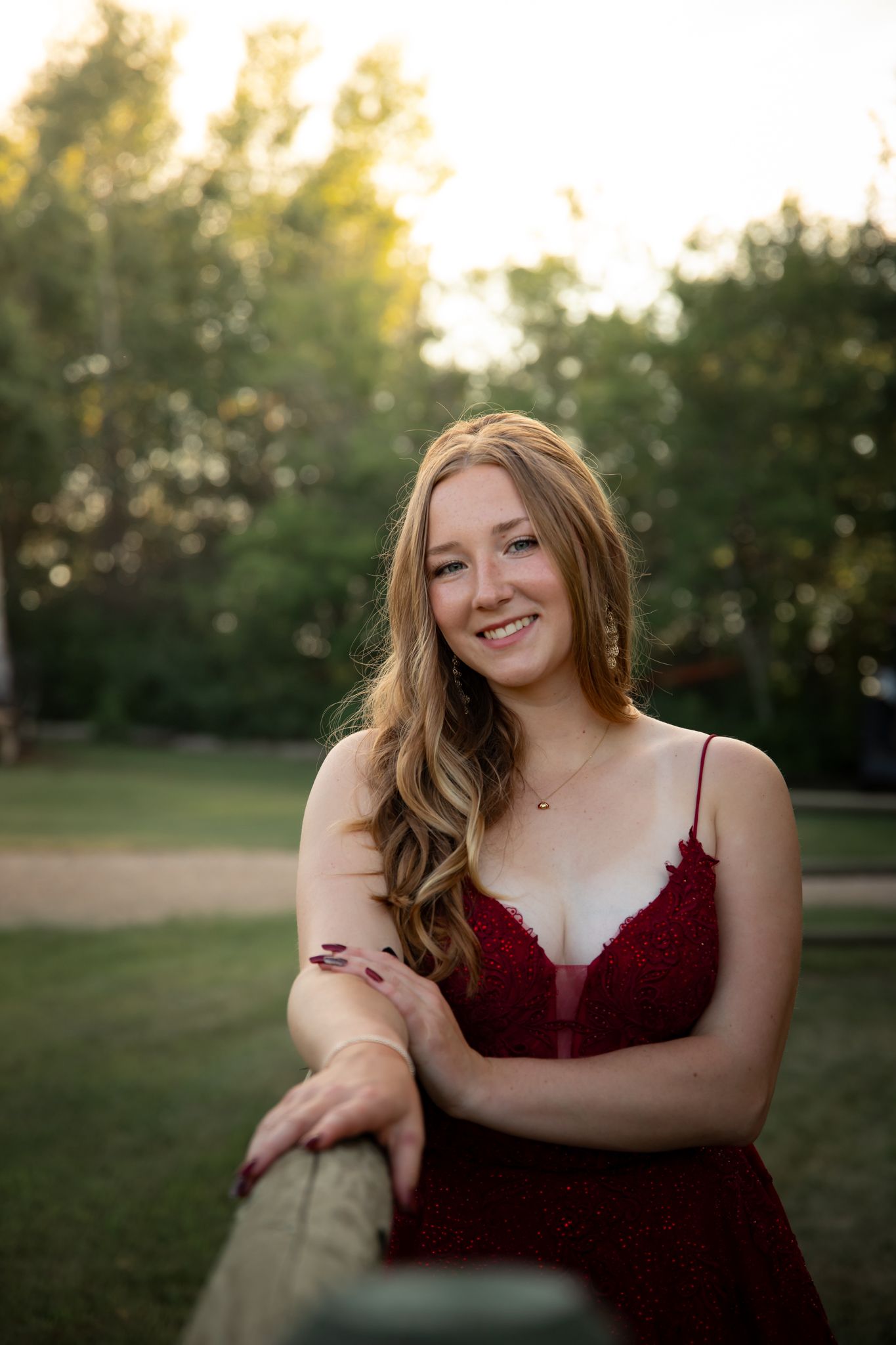 A person in a burgundy dress smiles while posing in a park setting at sunset.