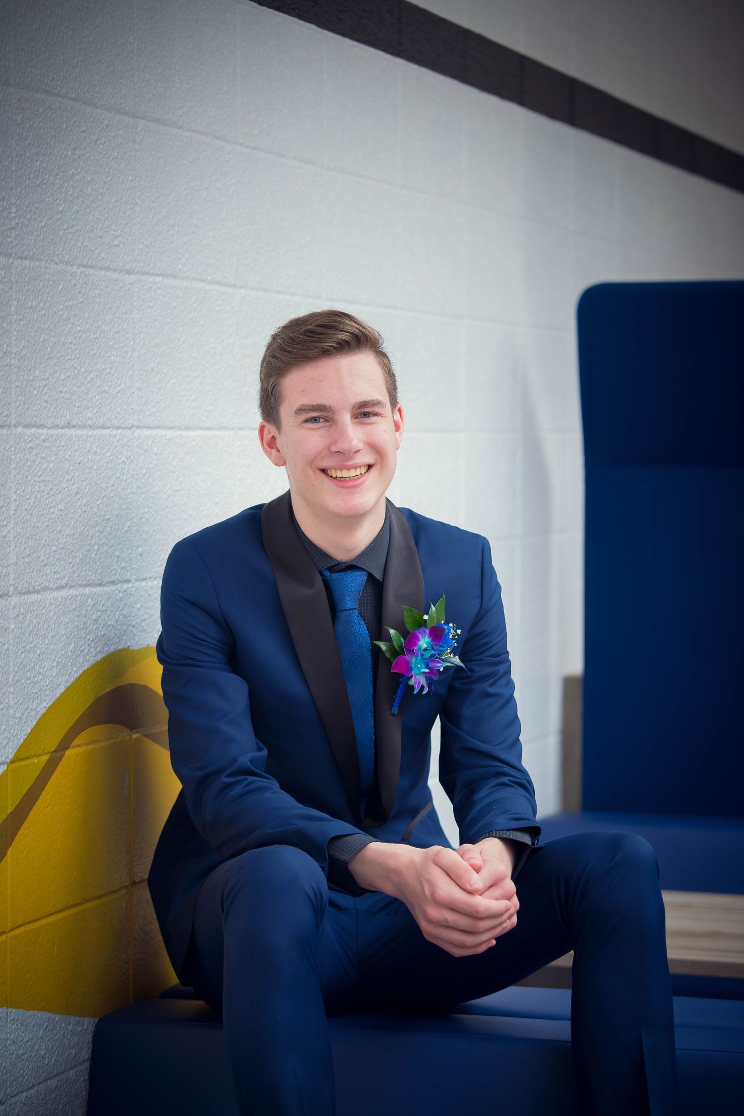 A young man in a navy blue suit sits casually on a blue bench against a white wall.