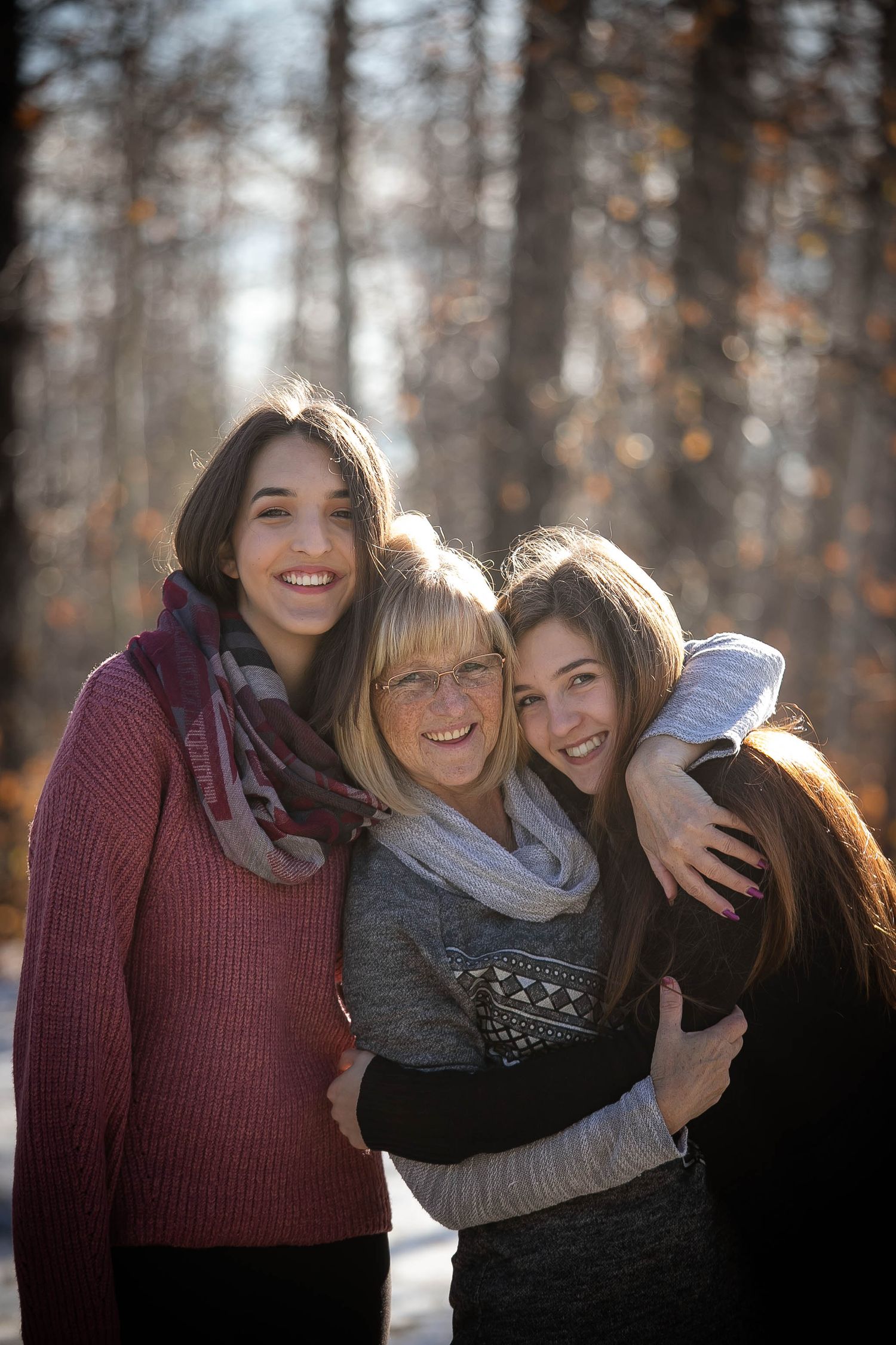 Three people share a warm embrace while backlit by sunlight in a forest setting.