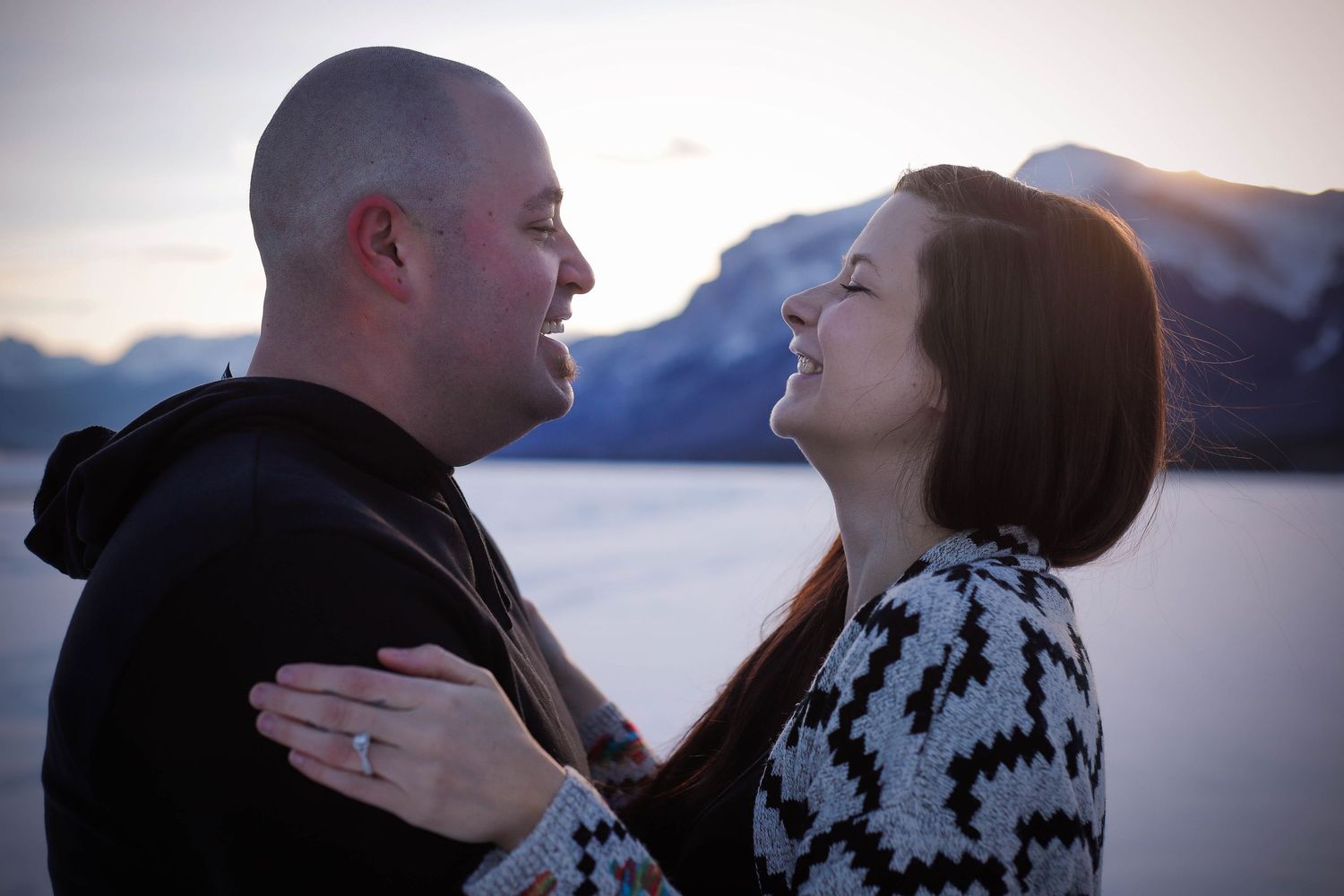 A romantic couple poses against a scenic mountain lake backdrop at sunset.