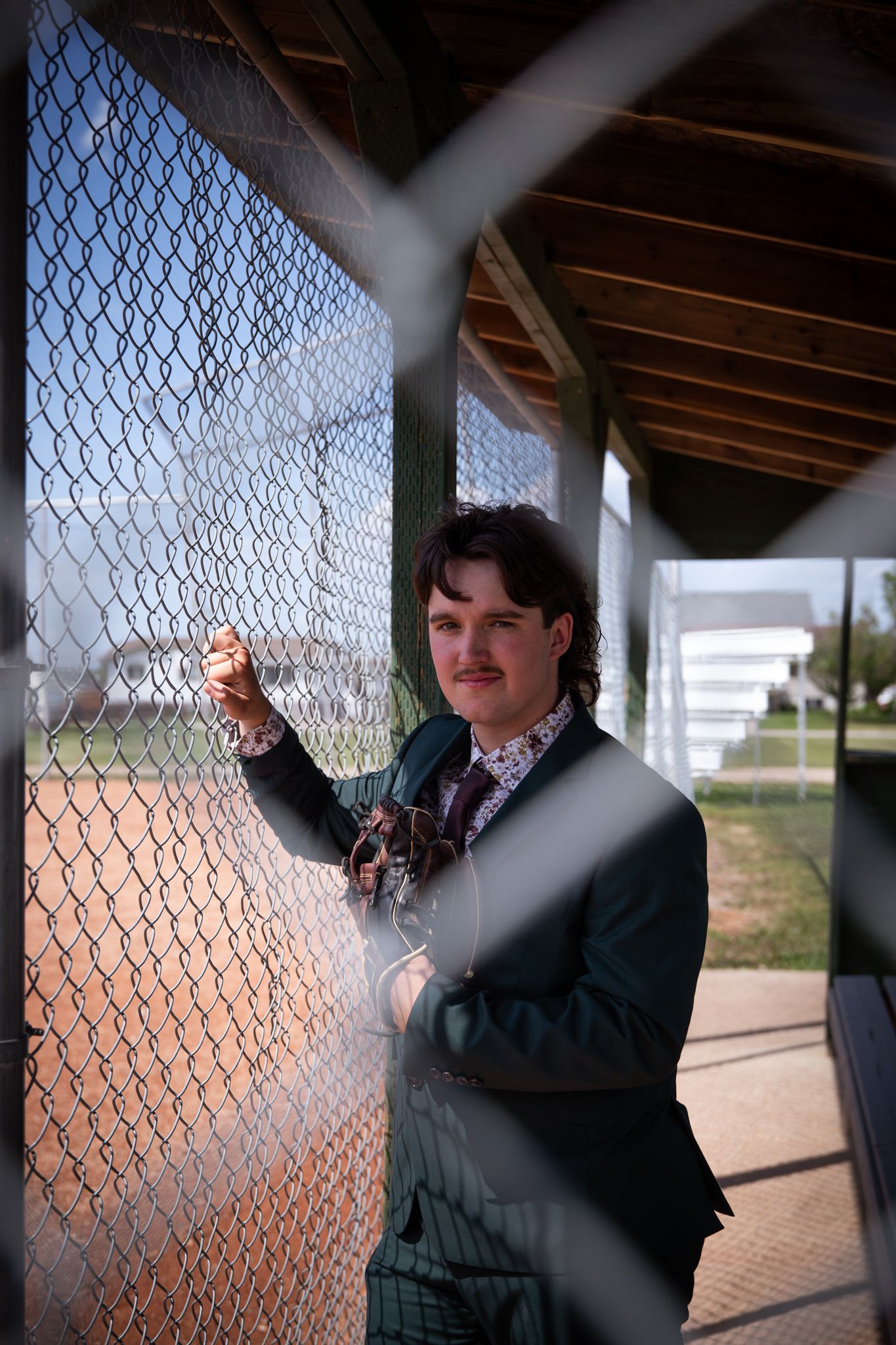 A person in formal attire stands by a chain-link fence in filtered sunlight.