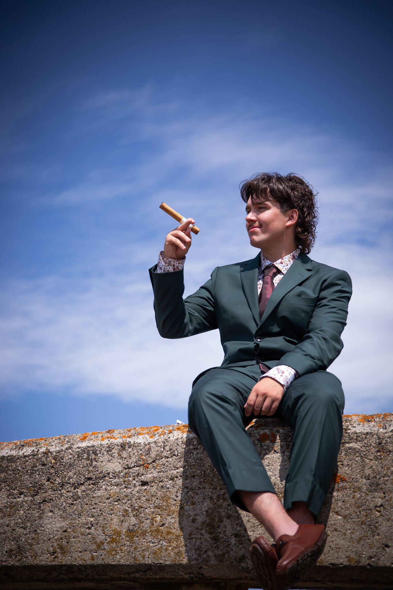 A suited figure sits casually on a concrete wall against a bright blue sky.