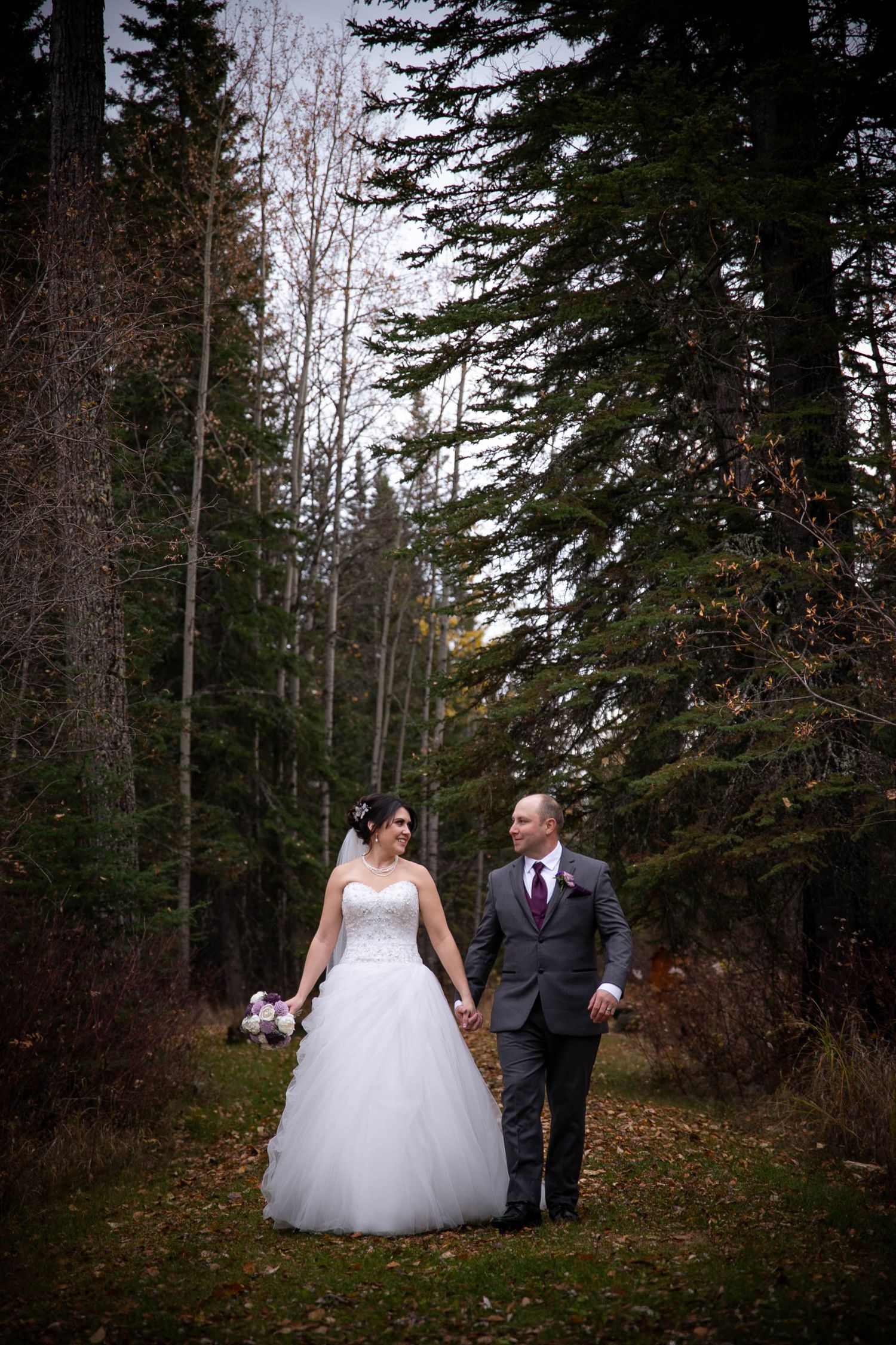 A couple walks through a wooded forest path in formal attire.