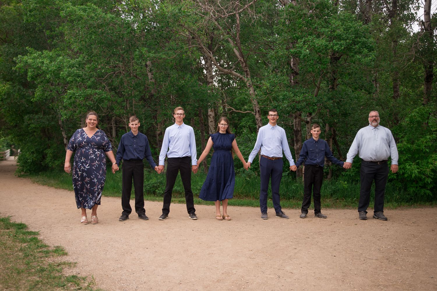 Group of people in formal attire standing in a line on a dirt path surrounded by trees.