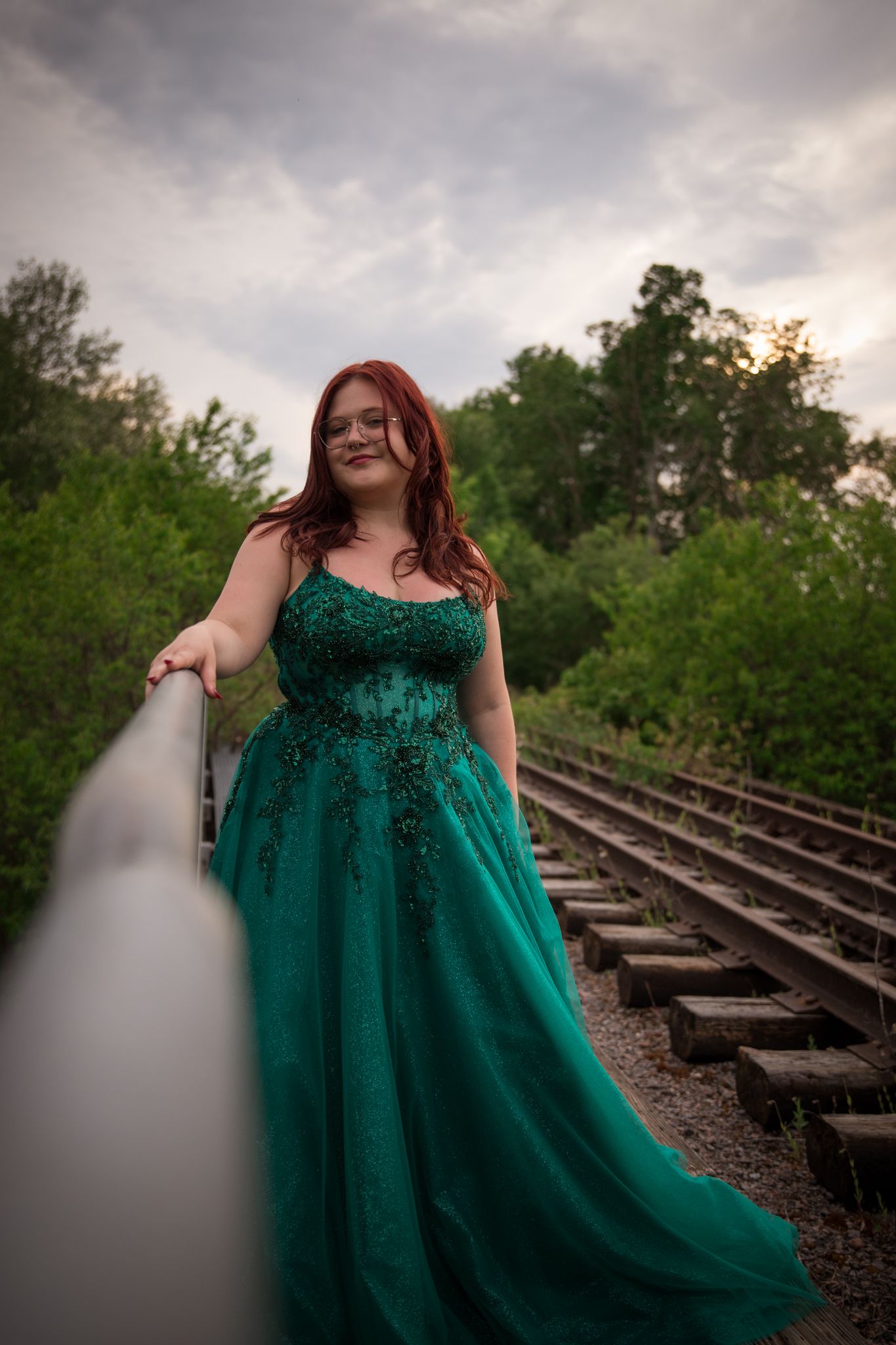 Emerald green formal gown photographed against dramatic cloudy sky on railroad tracks.