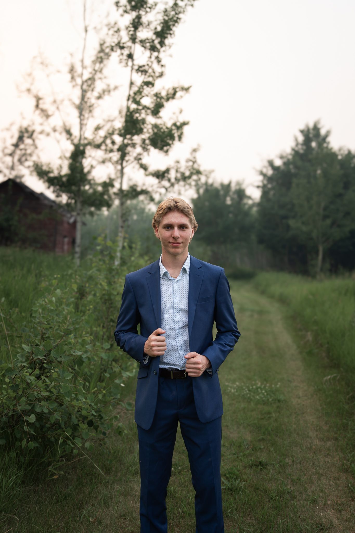 A person in business attire poses along a tree-lined rural path during golden hour.