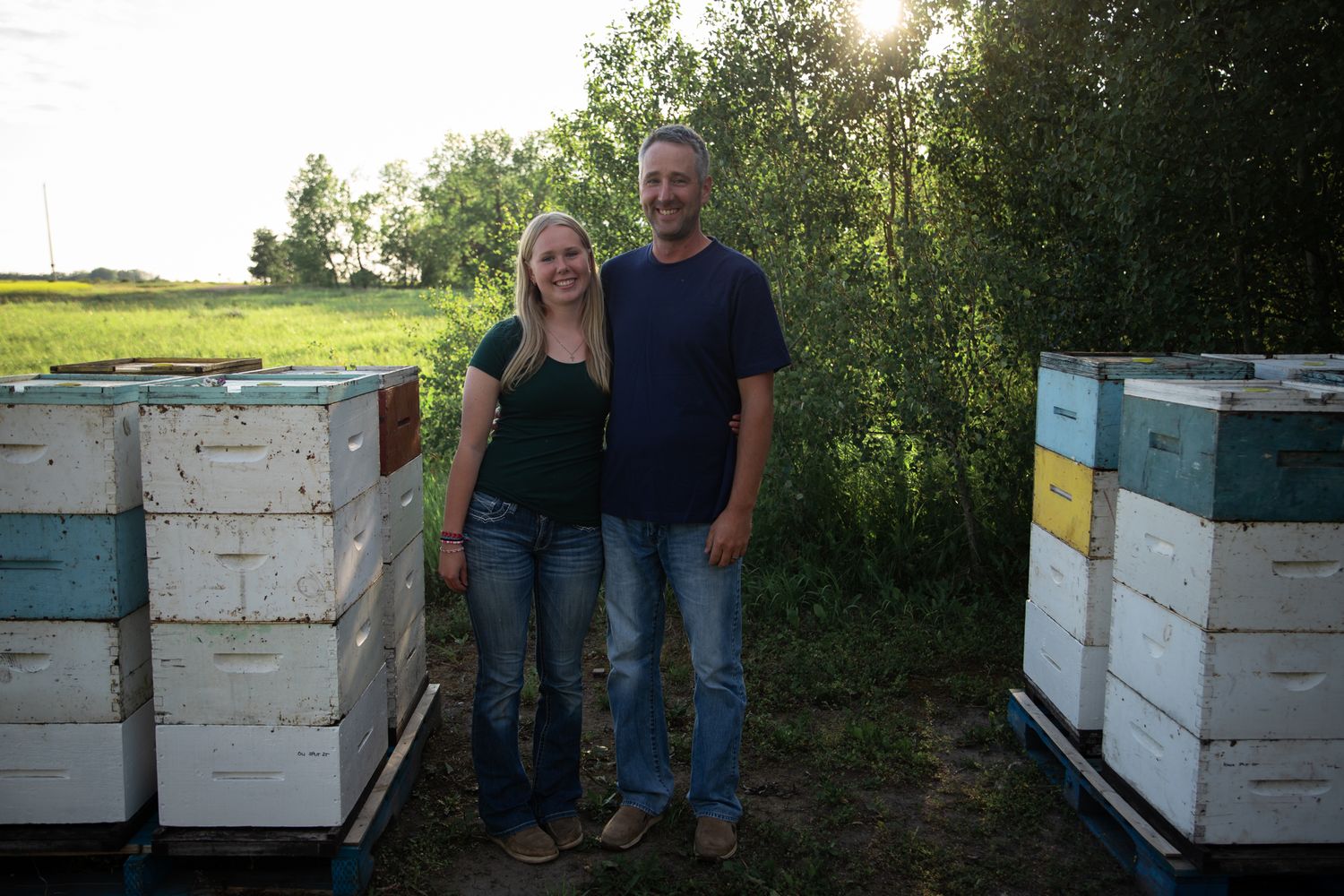 Two people stand between rows of white beehive boxes at dusk in a field.