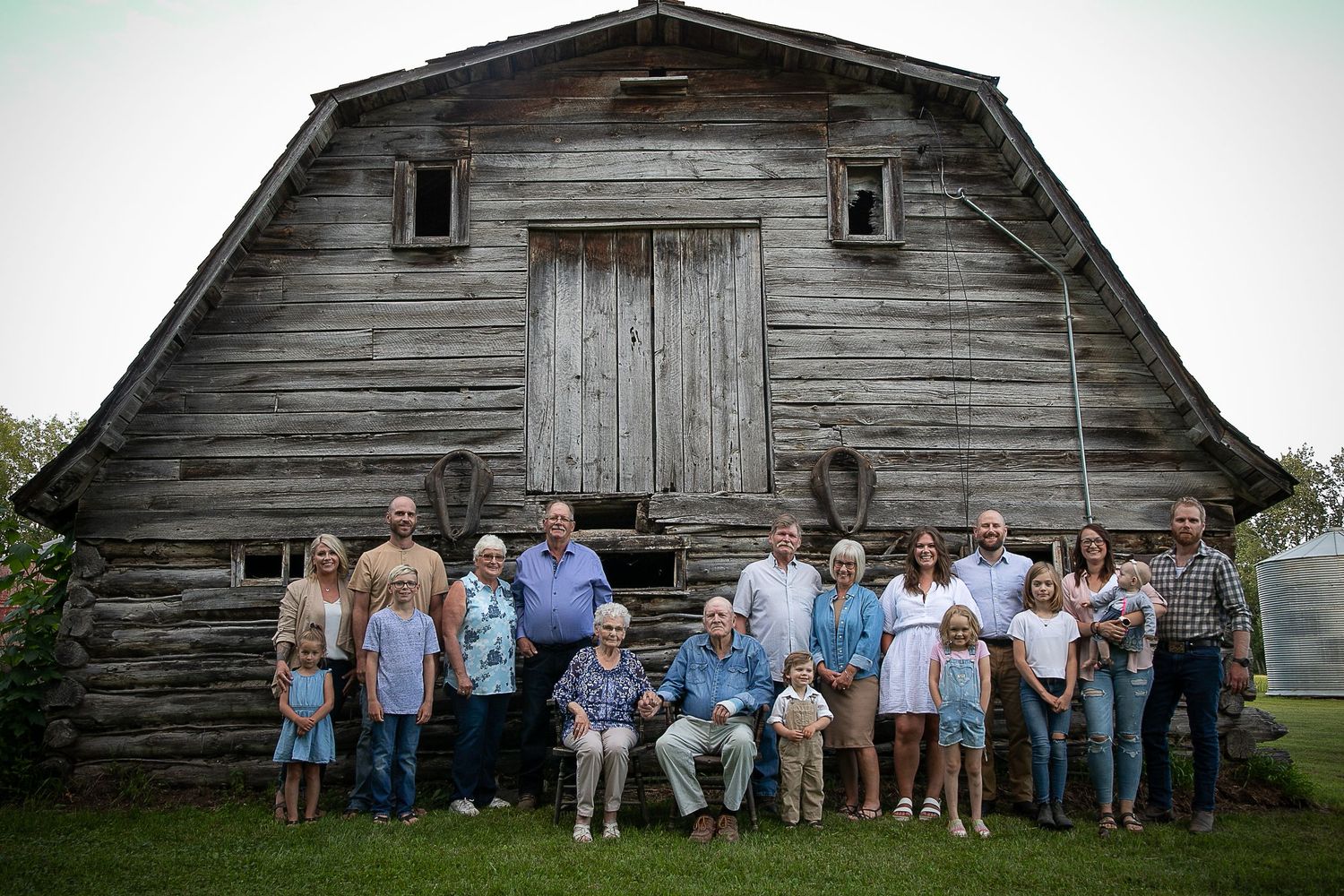 A large group poses in front of a weathered wooden barn with rustic architecture in a rural setting.