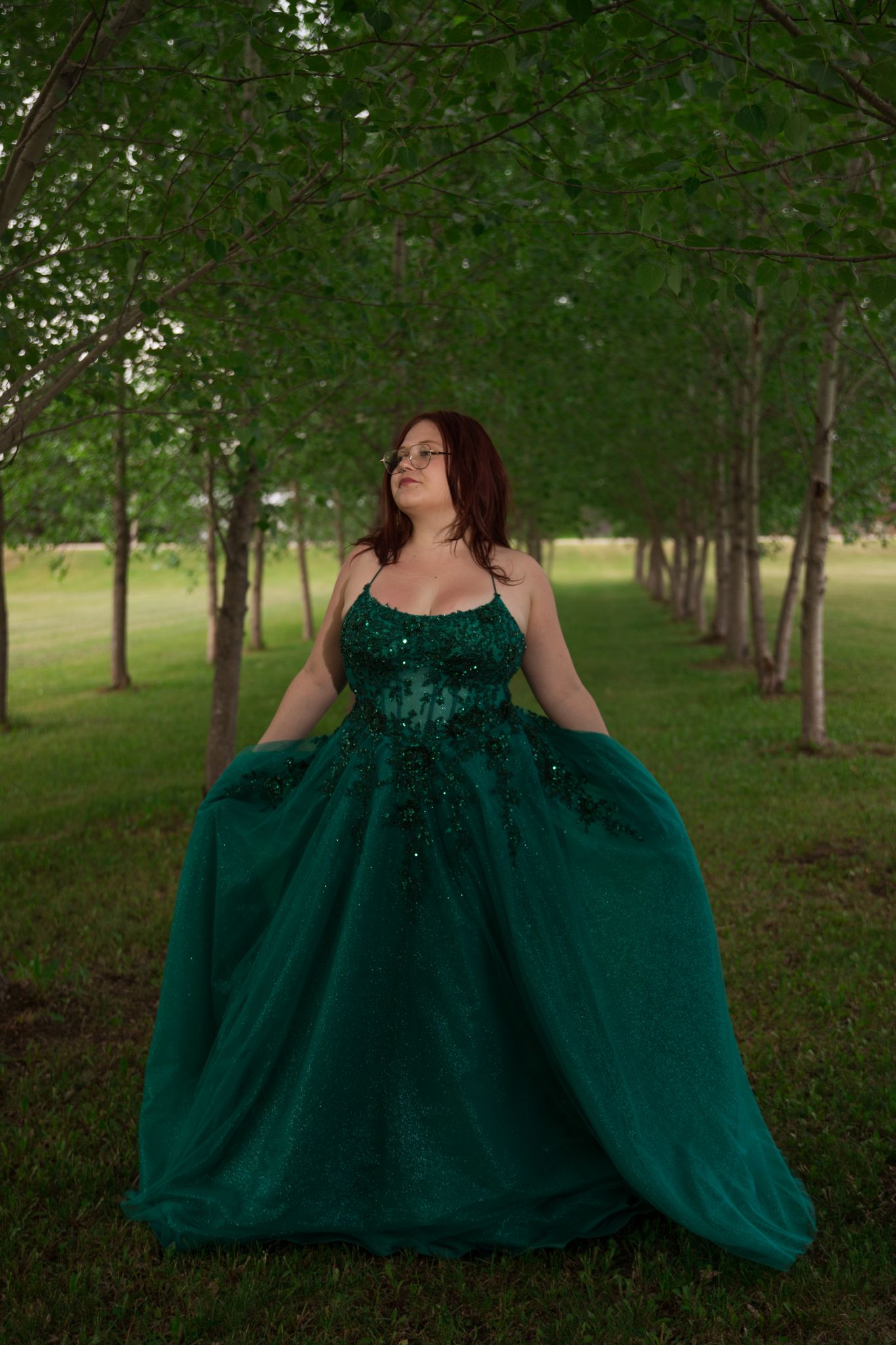 A flowing emerald green ballgown dress photographed in a scenic tree-lined outdoor setting.