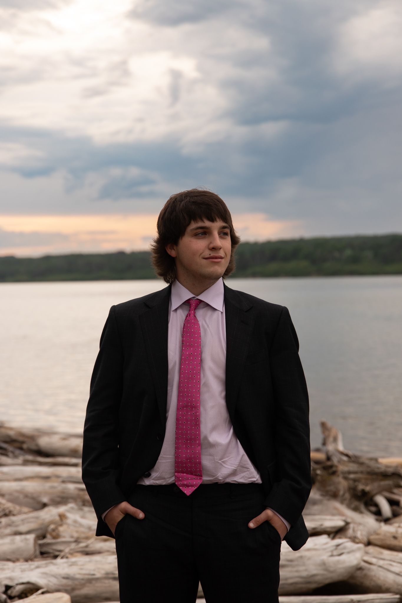 A person in a black suit with pink tie sits on driftwood by a lake at sunset.
