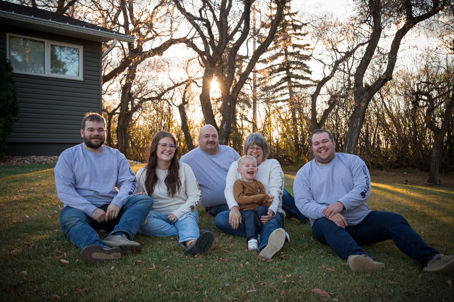 Family group sitting together on grass with trees and autumn foliage in background.