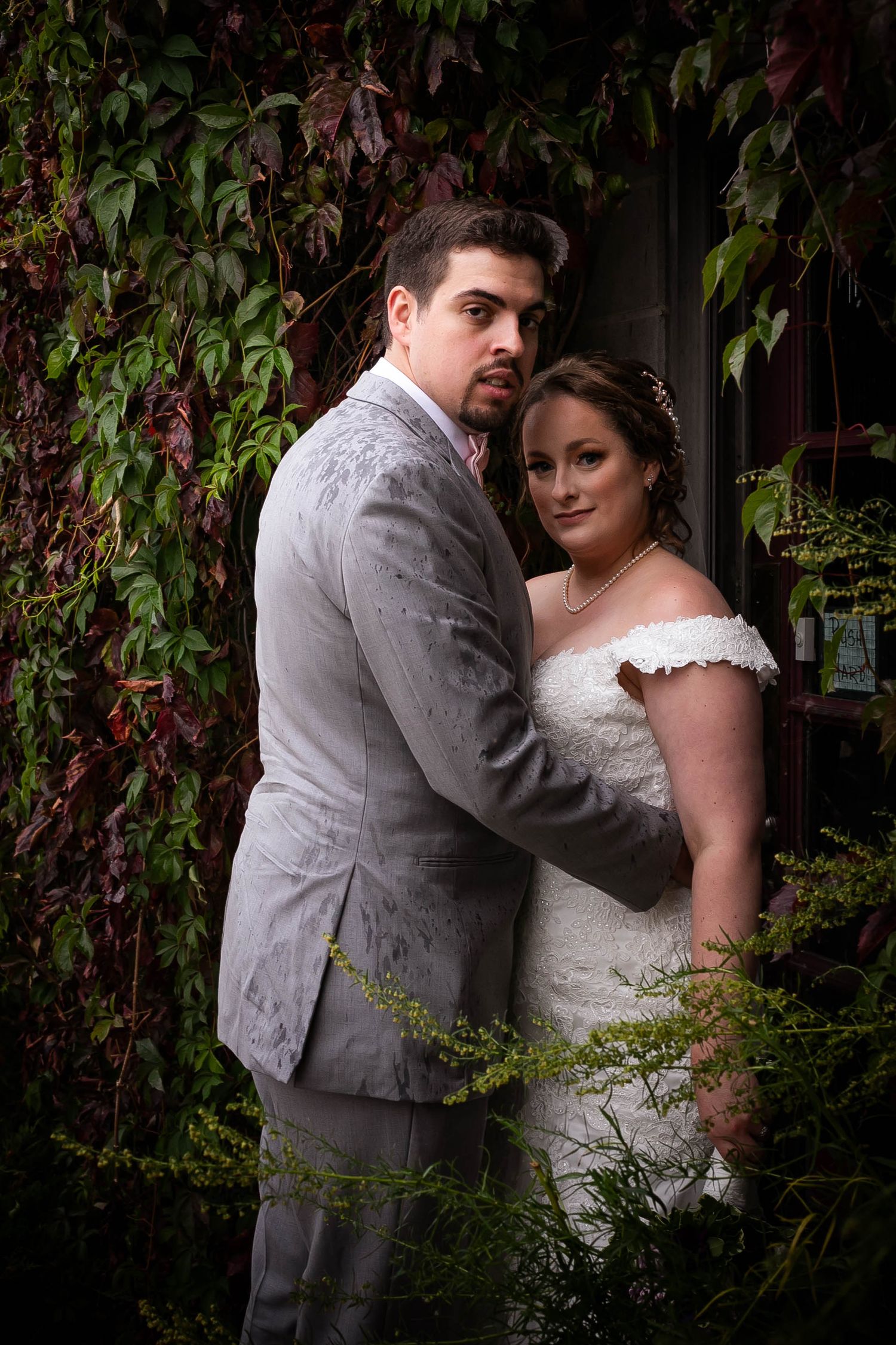 A romantic wedding couple poses against a vine-covered wall in an intimate garden setting.