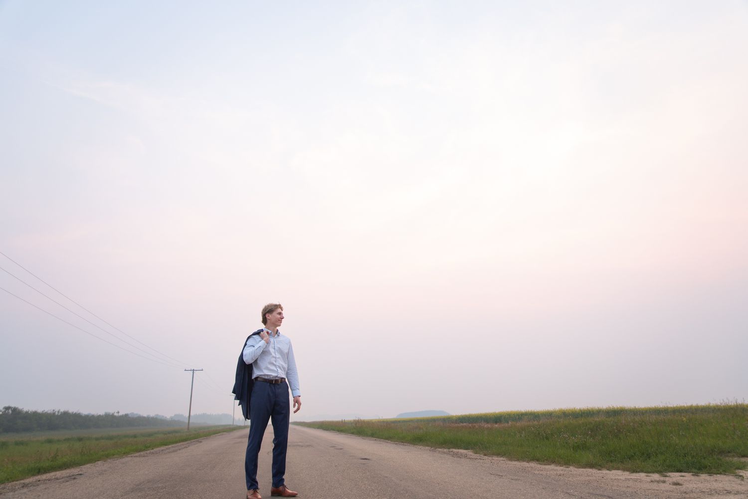 A solitary figure stands on an open rural road against a pale sky at dusk.