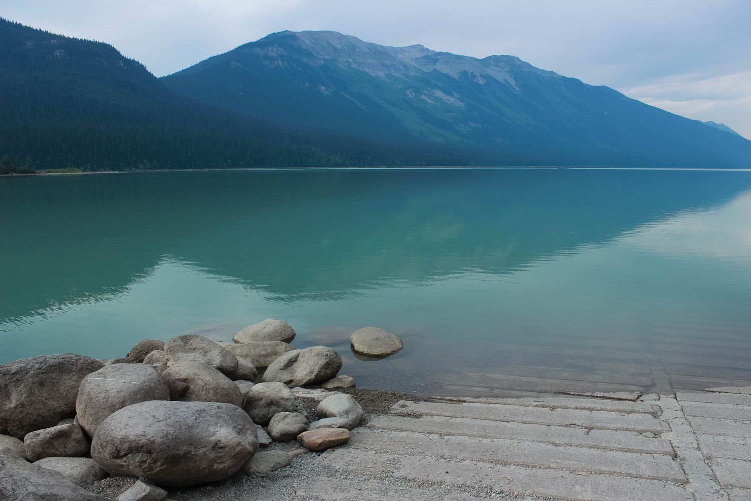 Tranquil turquoise lake with mountain reflections and rocky shoreline in foreground.