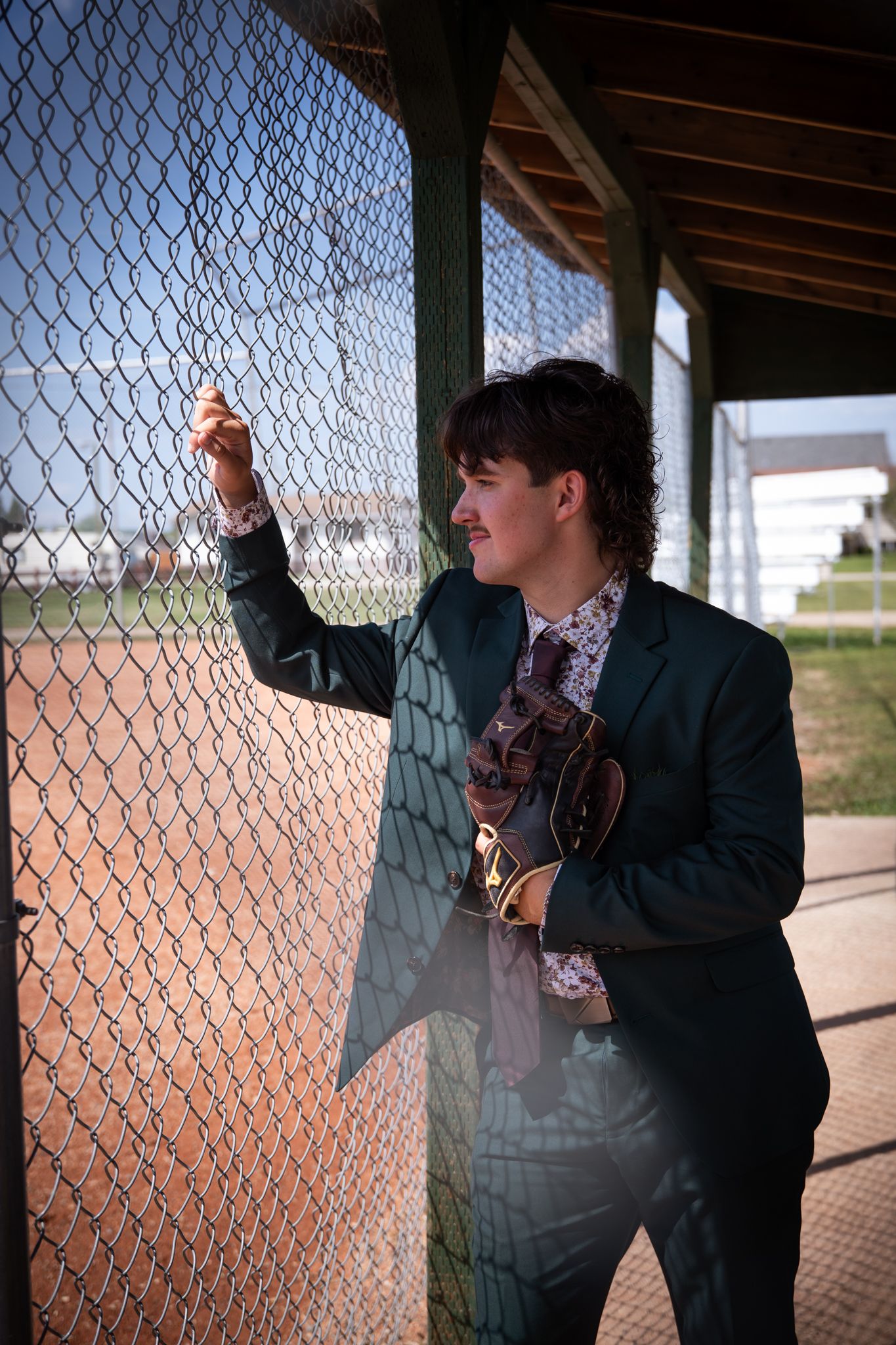 Someone in a dark suit poses playfully against a baseball field fence.