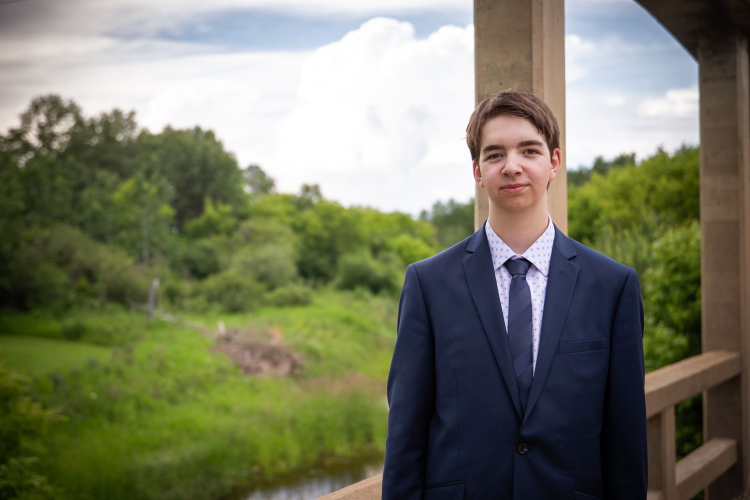 A person in formal attire stands on a rustic wooden bridge with trees in the background.