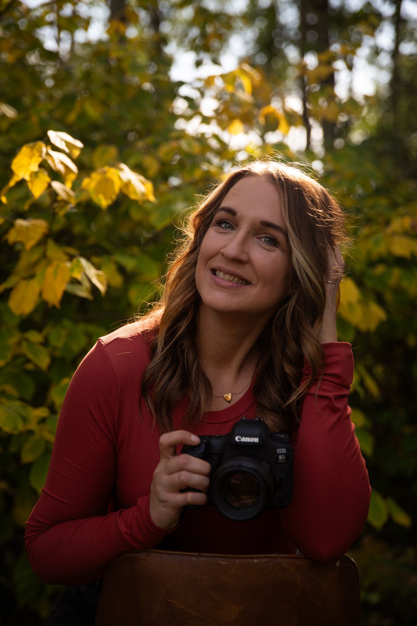 A photographer smiles while holding a Canon camera against autumn leaves in golden light.