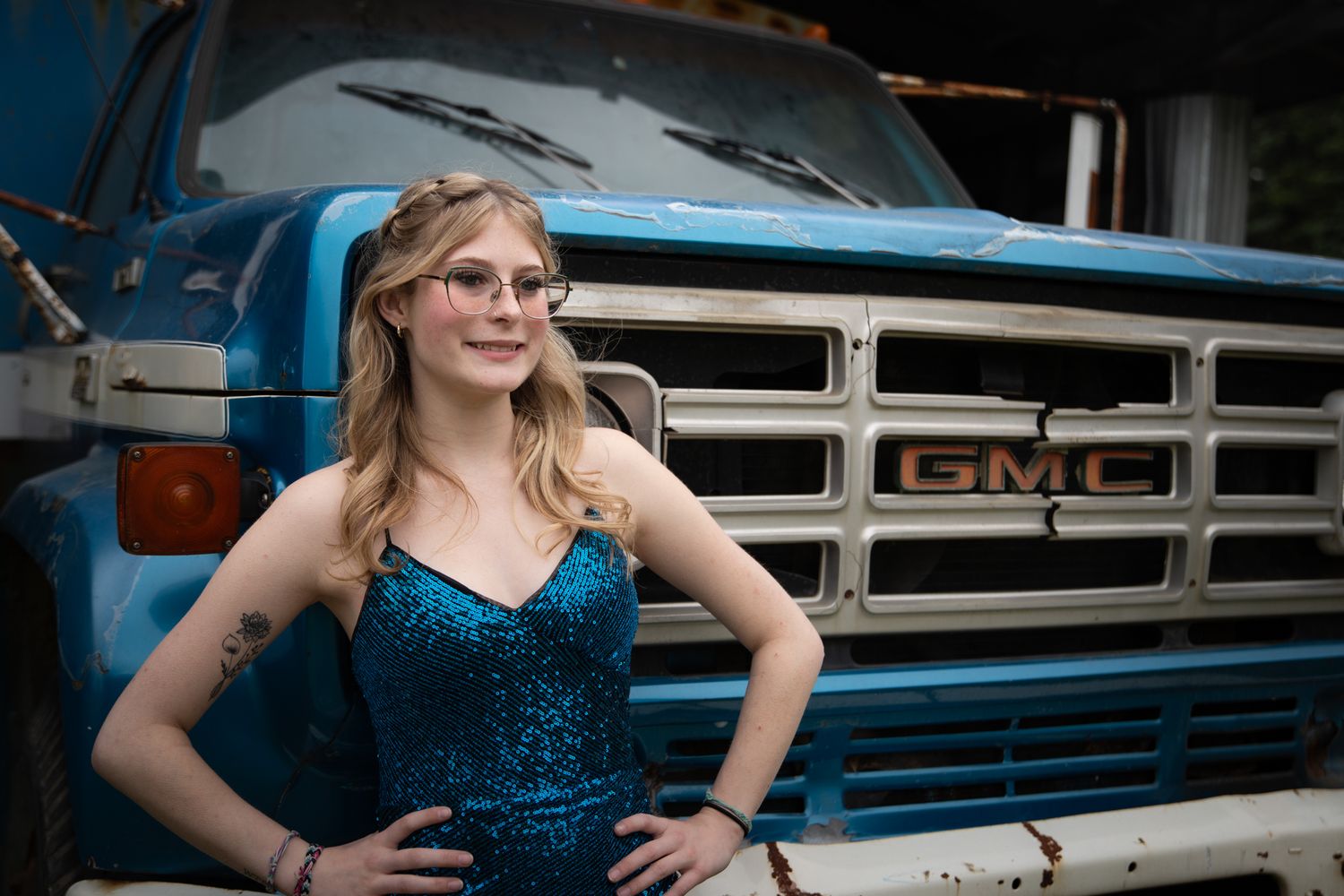 A sparkling blue sequin dress contrasts against vintage vehicles in an outdoor photoshoot setting.