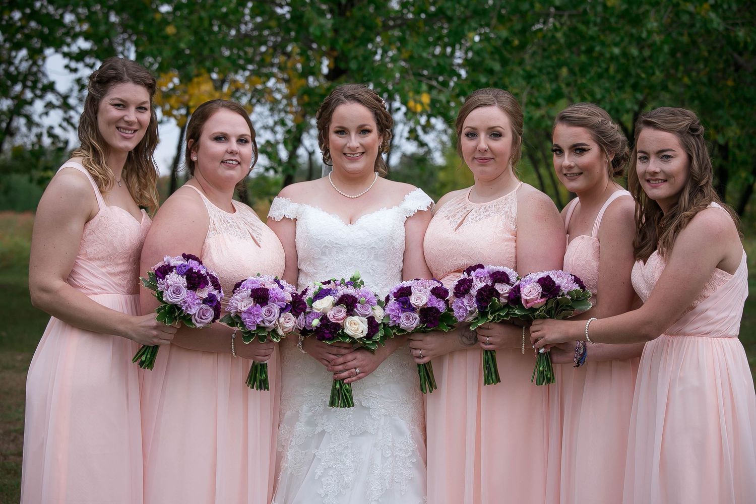 Bridesmaids in pink dresses hold purple floral bouquets while standing in a row.