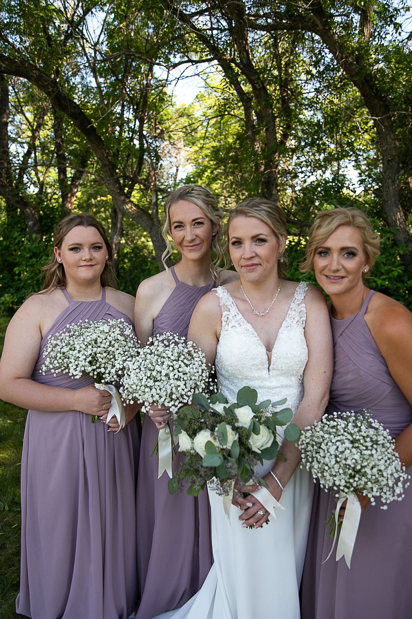 Wedding party in lavender dresses poses together holding baby's breath bouquets outdoors.