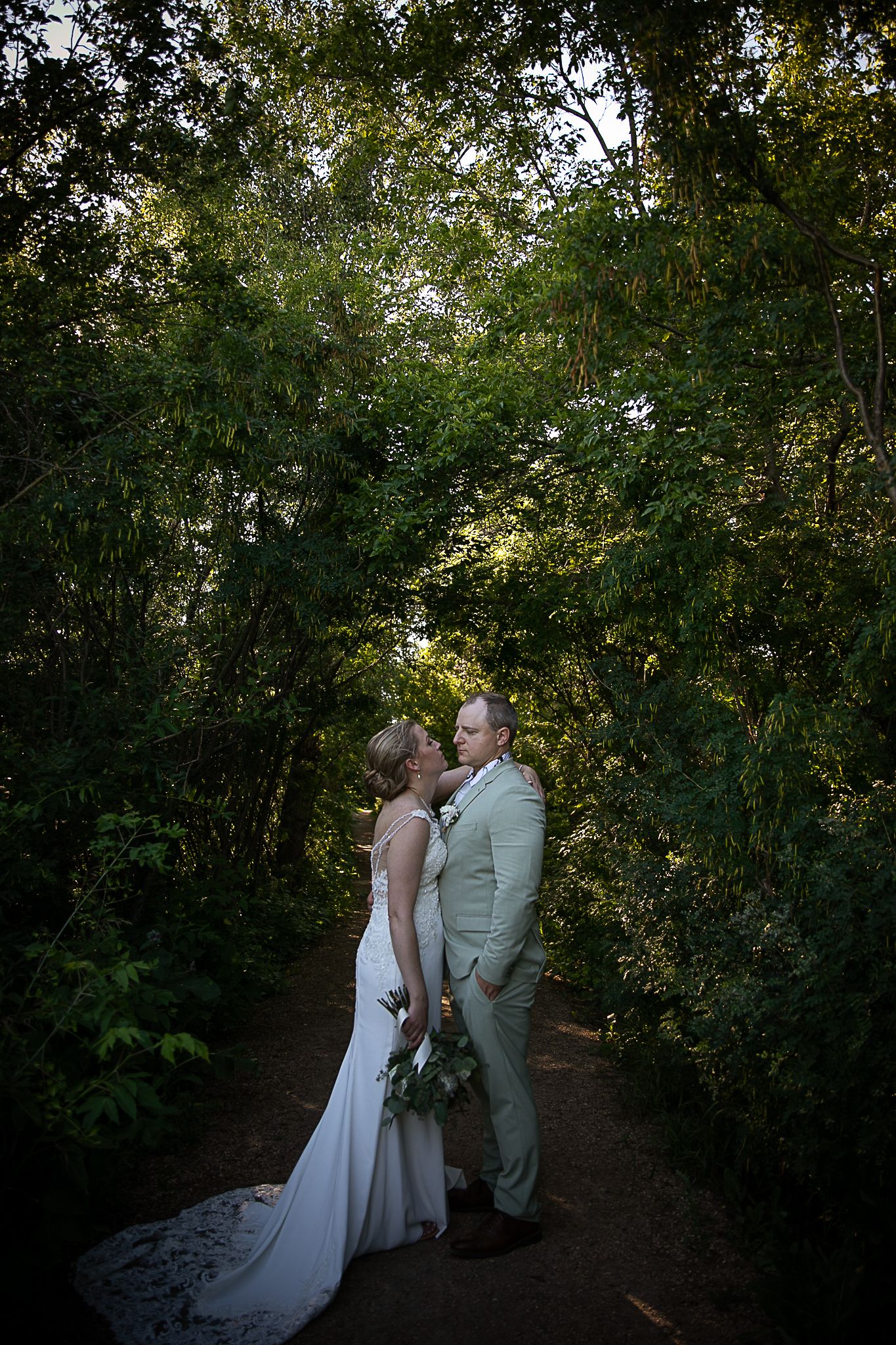 A romantic wedding couple embraces on a forest path surrounded by lush greenery at sunset.
