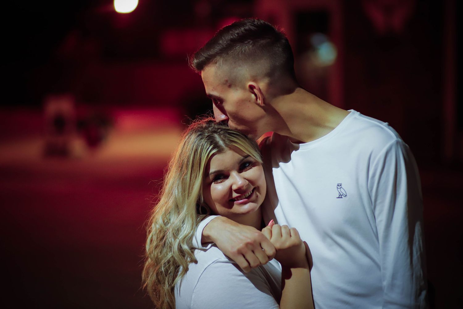 A couple shares an intimate moment against a dark red background with dramatic lighting.