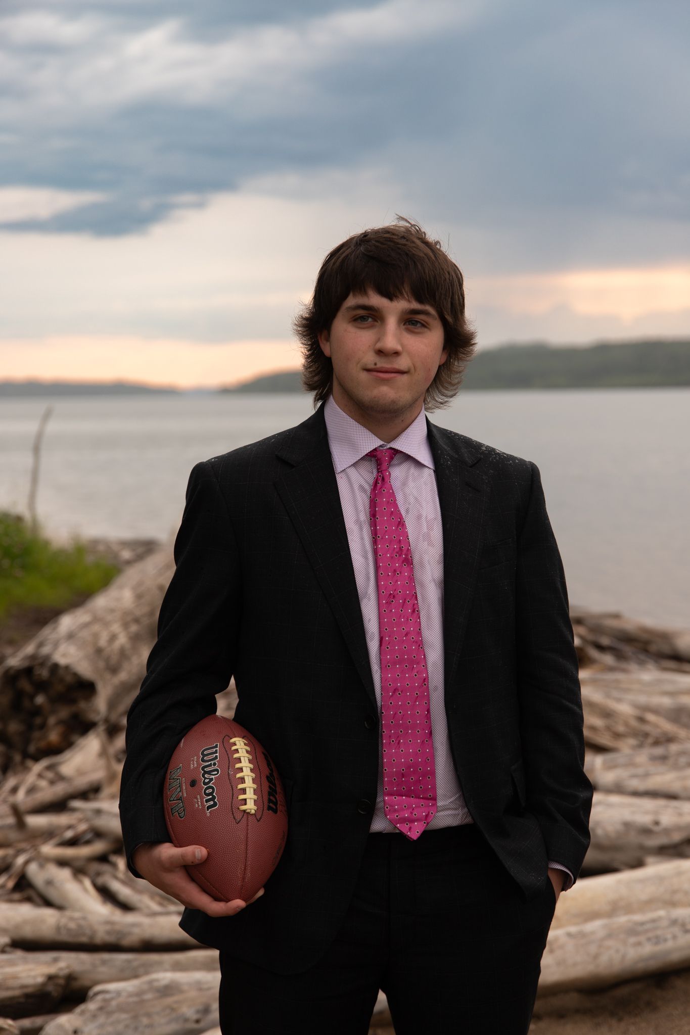 A person in formal wear poses with a football against a lakeside backdrop at dusk.