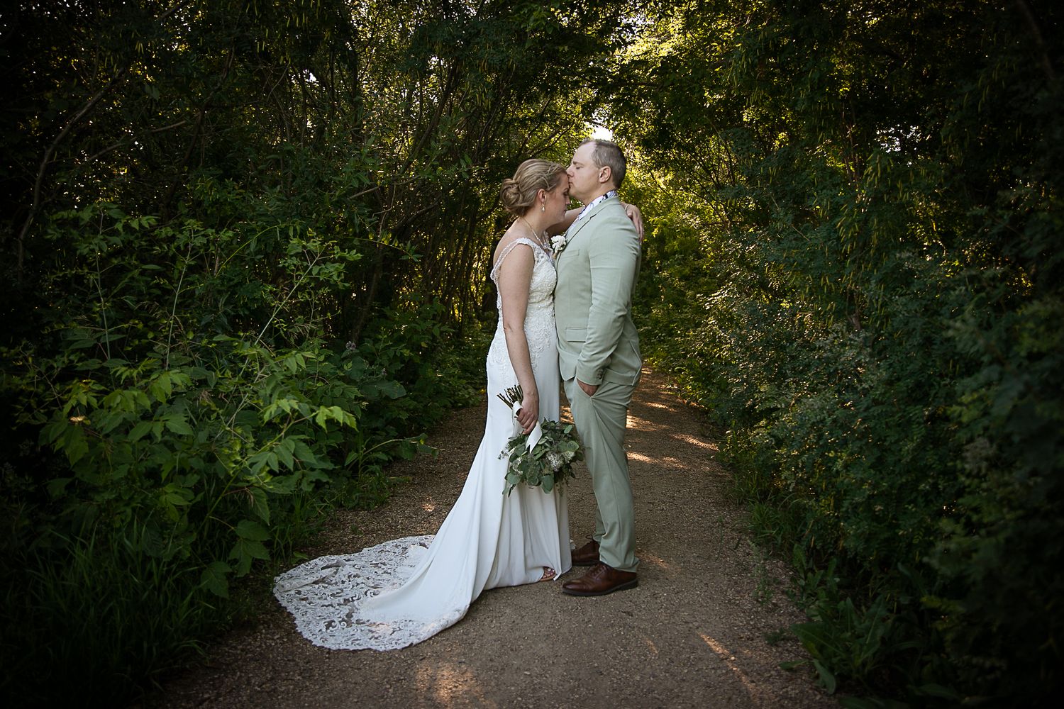 A couple in wedding attire stands together on a tree-lined path during their outdoor photoshoot.