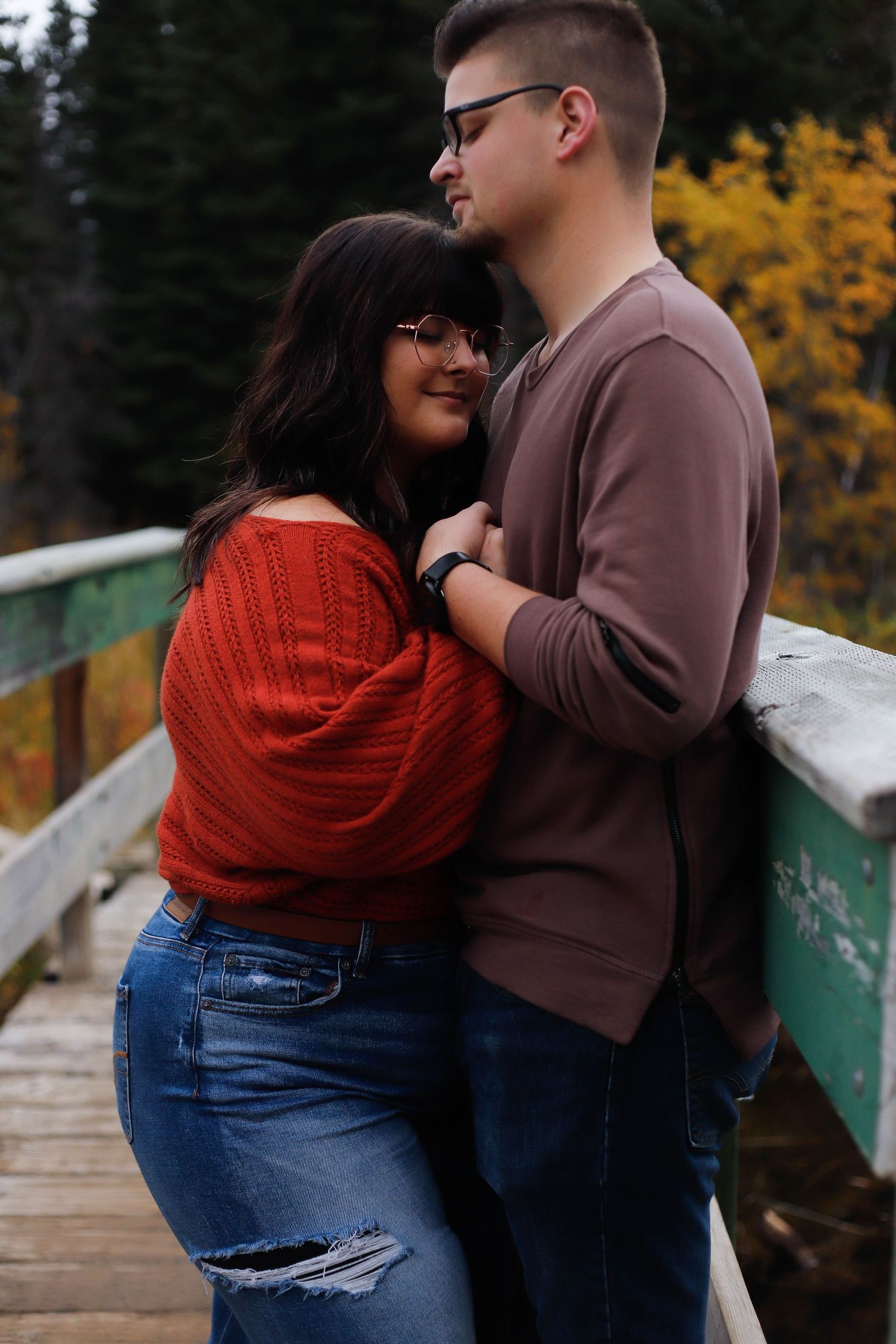 A couple shares an intimate moment on a bridge during autumn, with vibrant fall colors surrounding them.