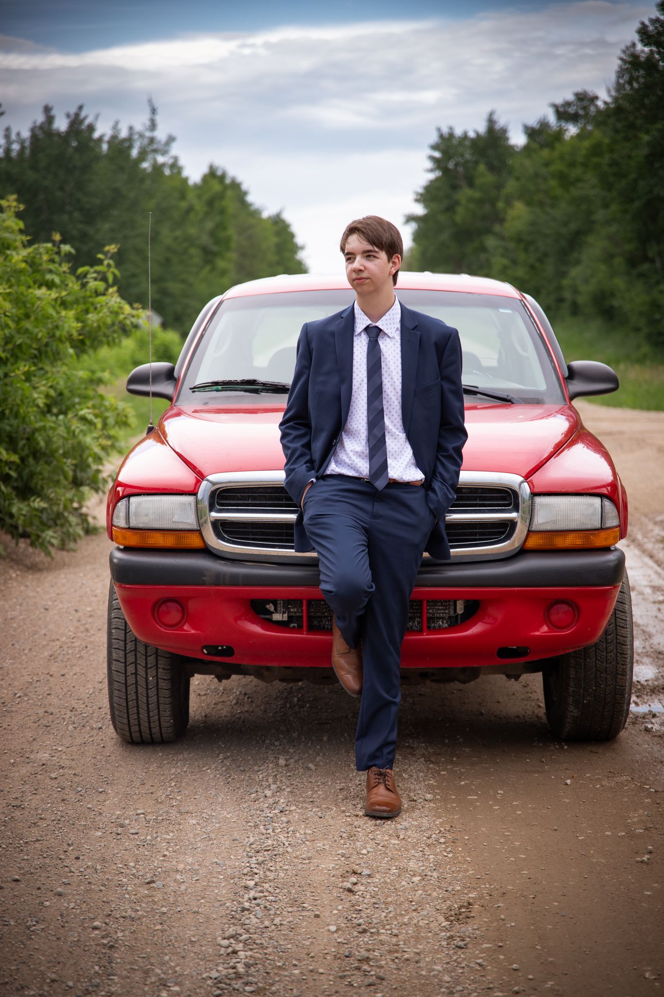 A person in formal wear poses with a classic red sports car on a gravel path.
