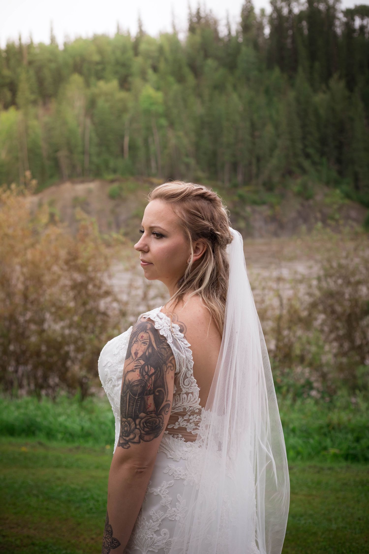A bride in a lace wedding dress with a flowing veil poses against a natural forest backdrop.
