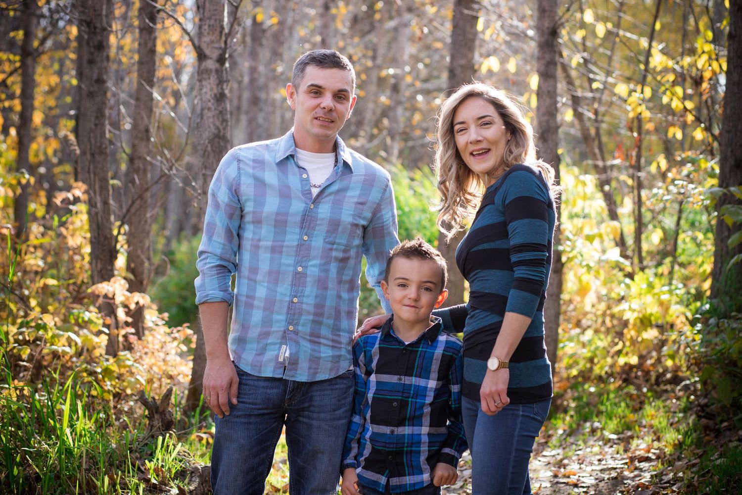 Family of three in matching plaid shirts pose together in a sunlit forest.