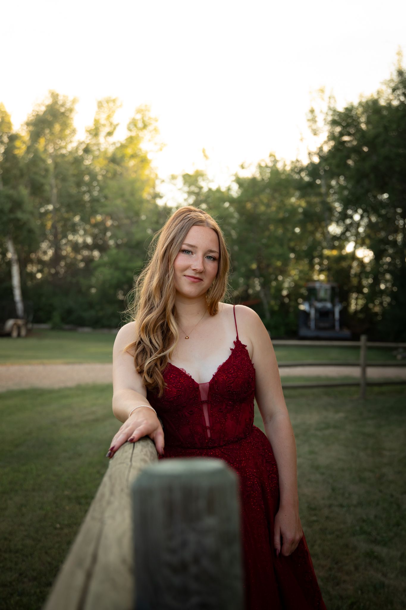 A person in a burgundy dress leans against a fence post in a park setting.