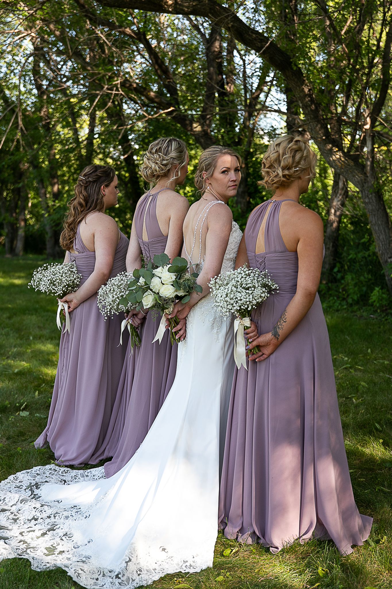 Bridesmaids in lavender dresses stand with bride holding white floral bouquets in garden setting.