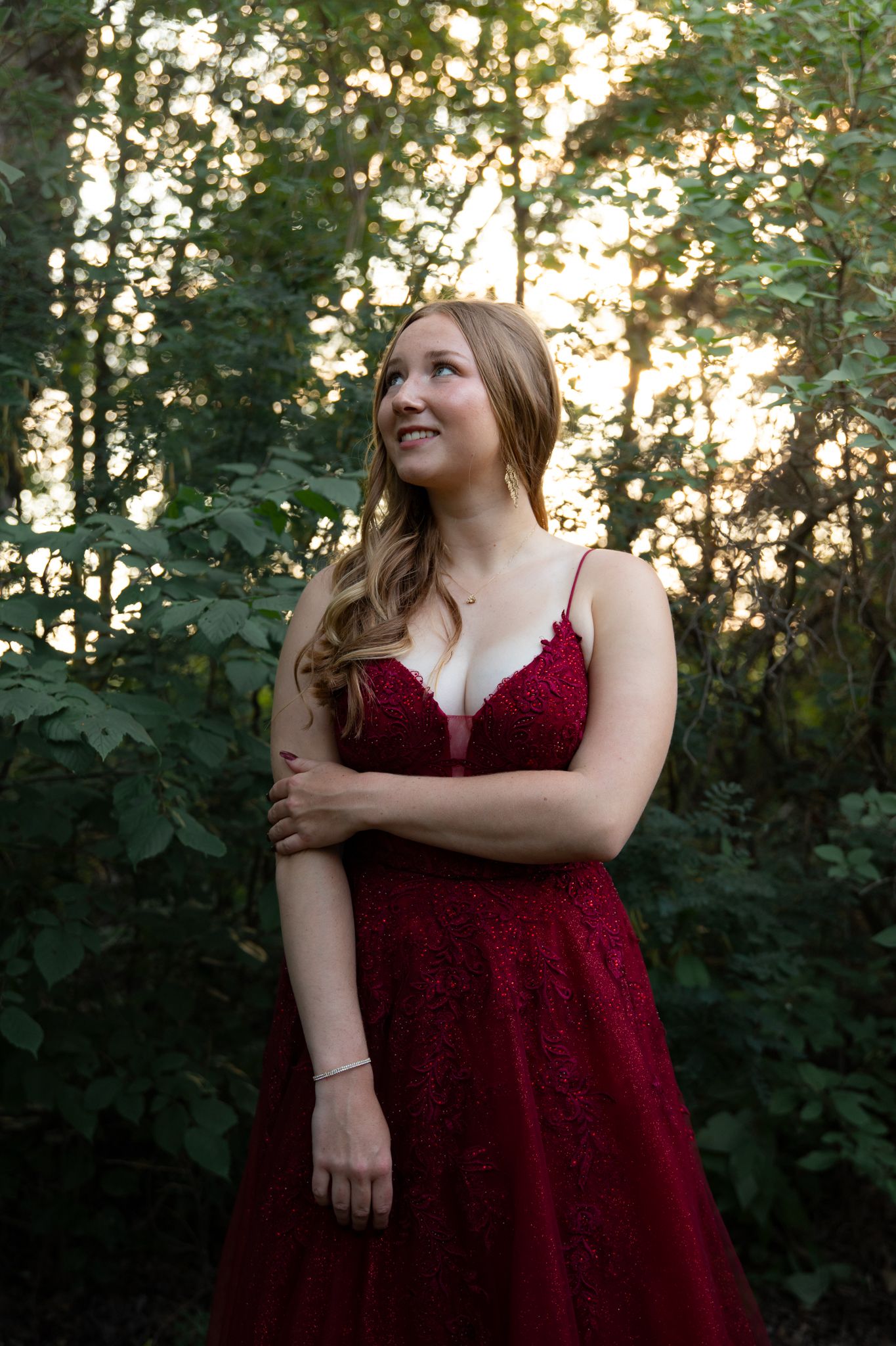 A burgundy lace formal dress photographed in a wooded setting during golden hour.