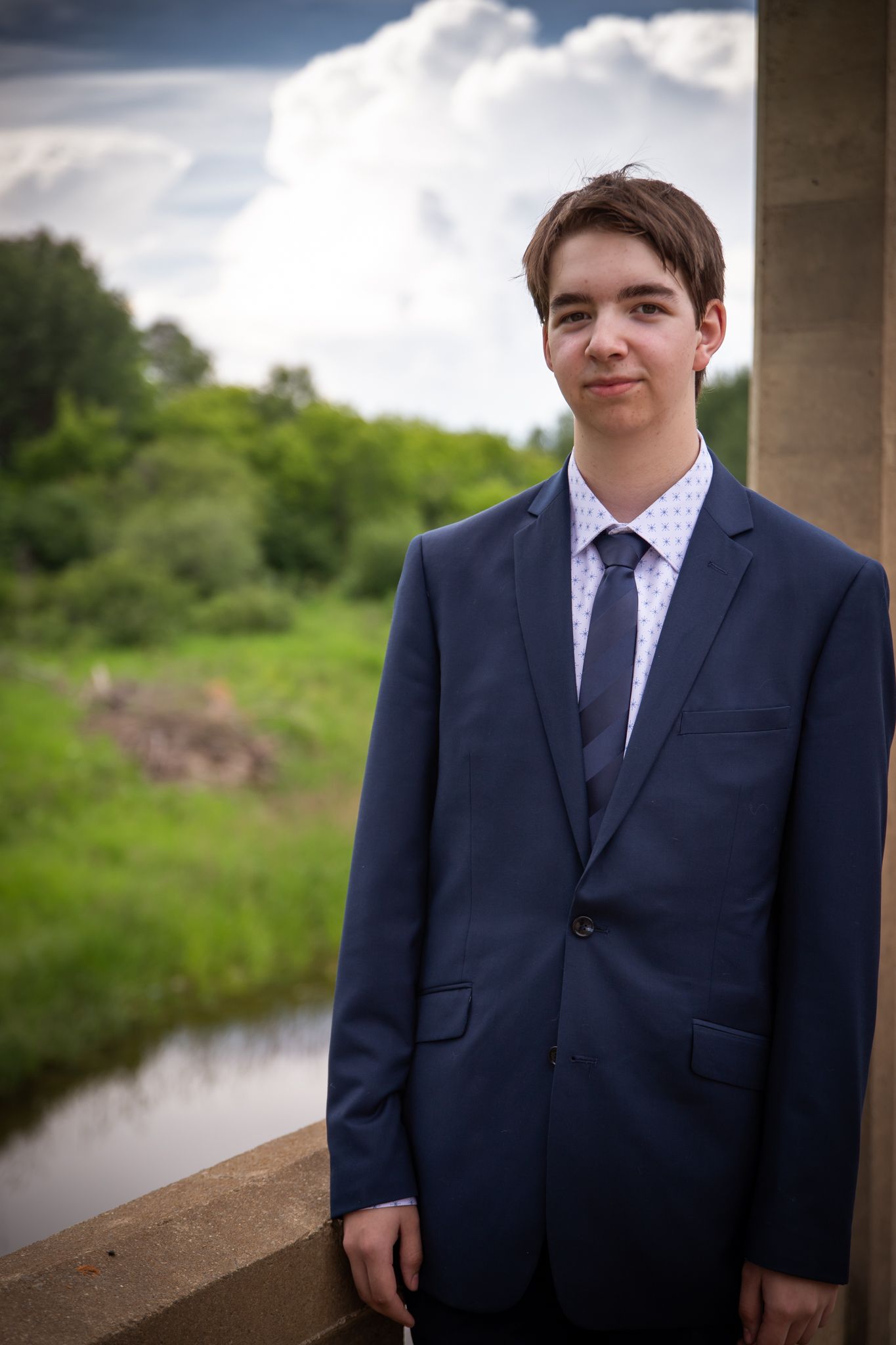 A person in a navy suit poses near a waterway with cloudy skies overhead.