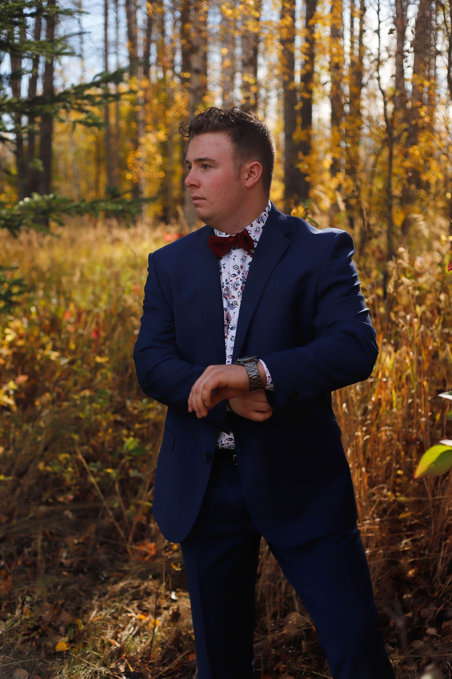 Person in navy suit poses thoughtfully among golden autumn trees.