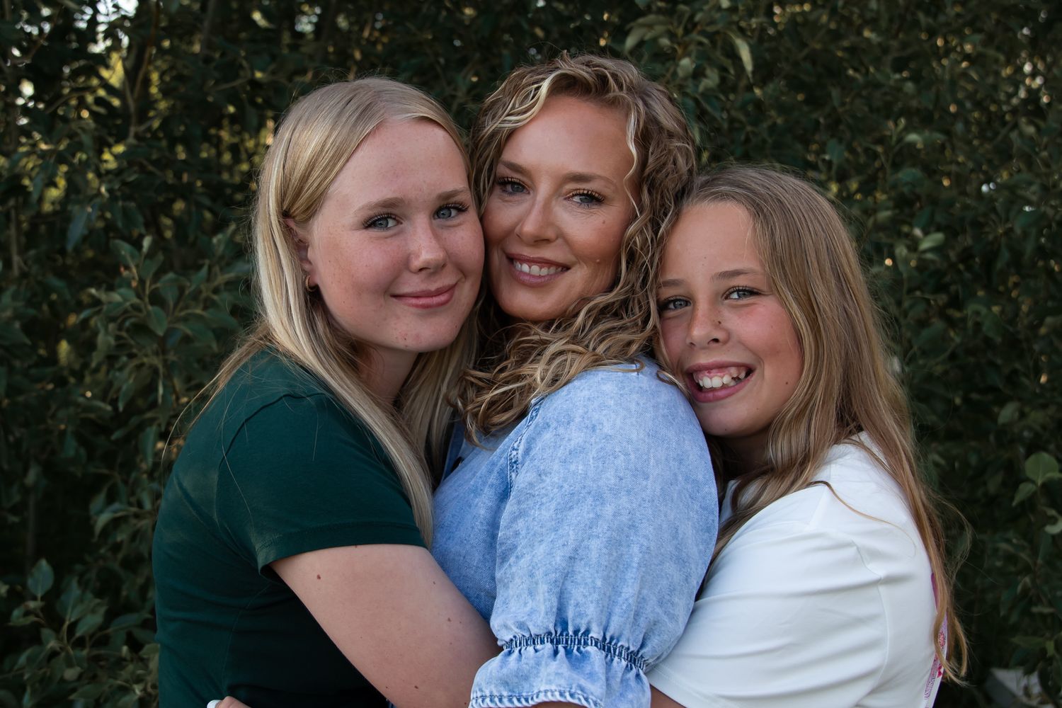 Three people embrace and smile together against a backdrop of green foliage.