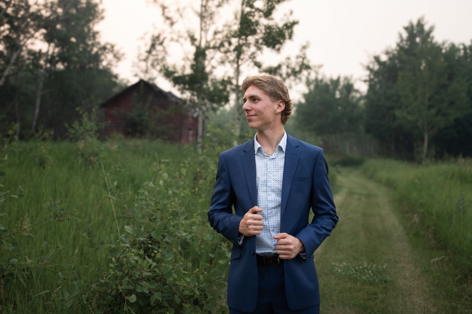 A person in a navy suit stands on a grassy path surrounded by lush greenery at sunset.
