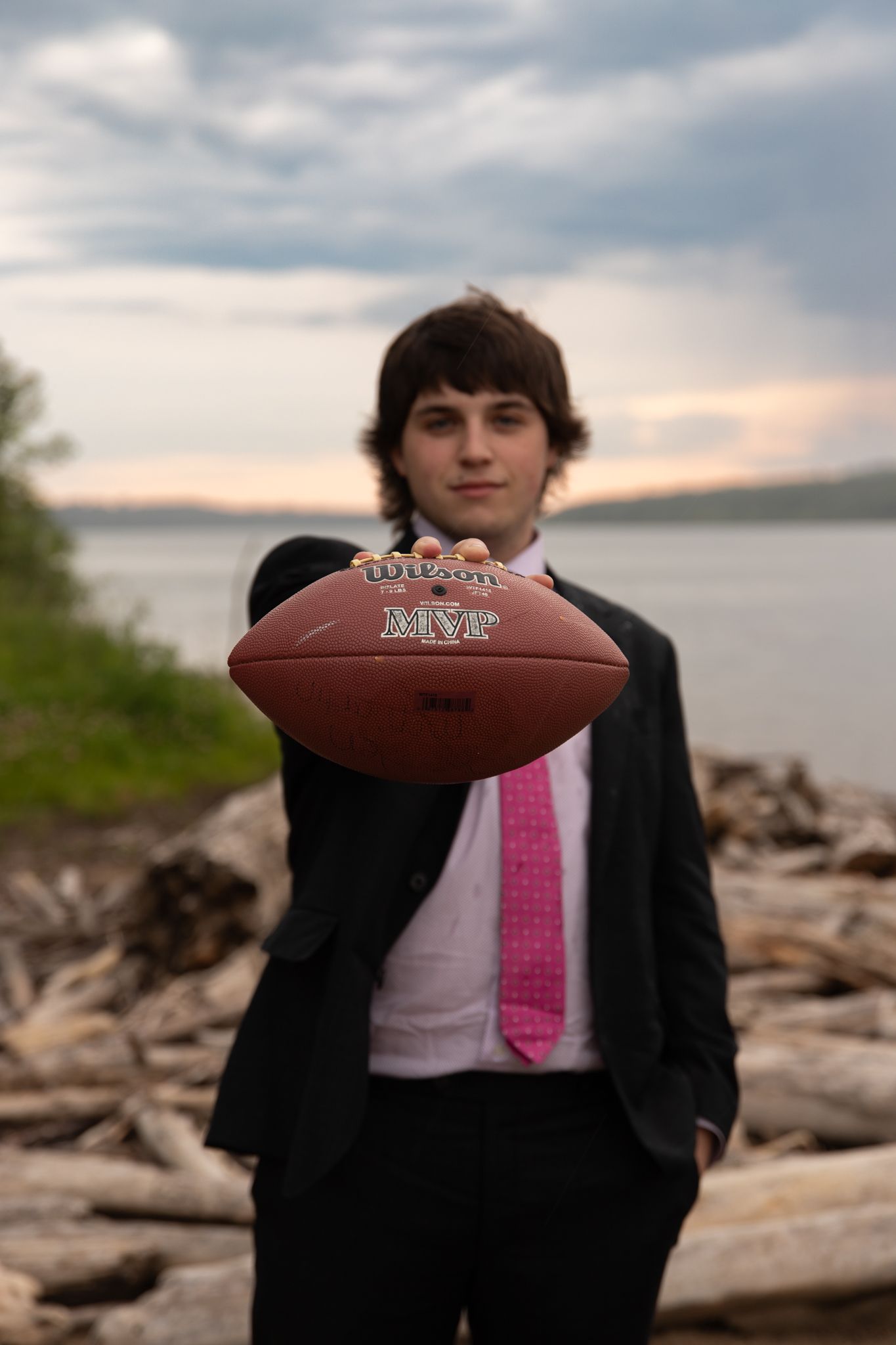 A person in formal attire holds a football while posing by driftwood on a beach.