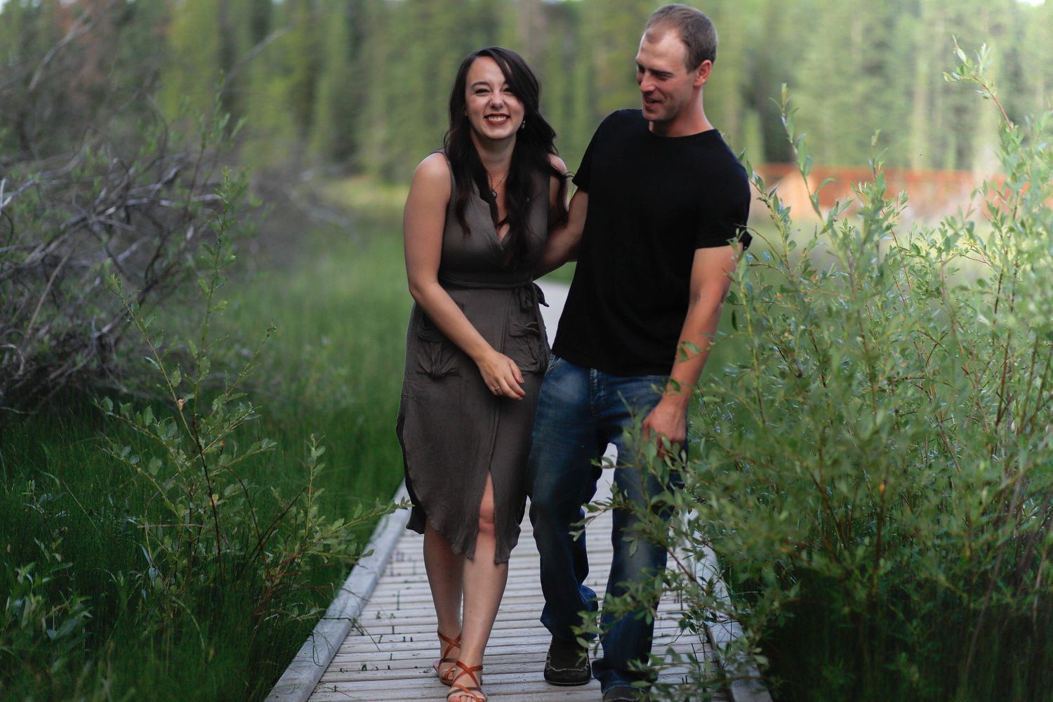 A joyful couple laughs together while walking on a wooden boardwalk surrounded by greenery.