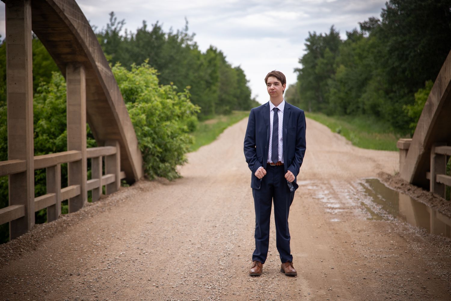 A figure in a dark suit stands on a dirt path surrounded by green foliage.