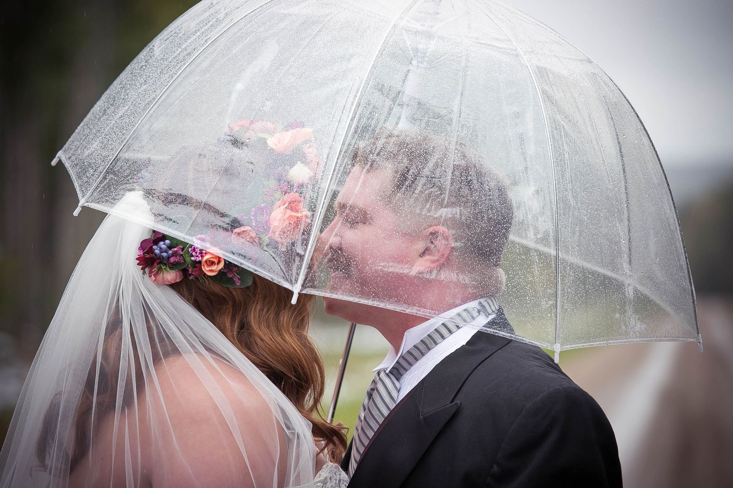 A clear umbrella frames a romantic wedding portrait in soft lighting.