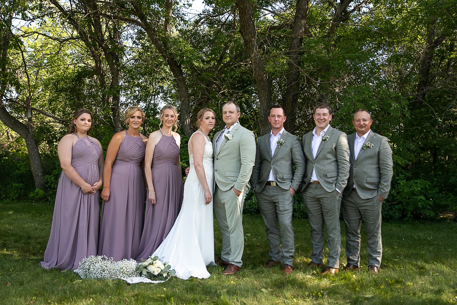 Wedding party in grey suits and lavender dresses poses together on grassy lawn.