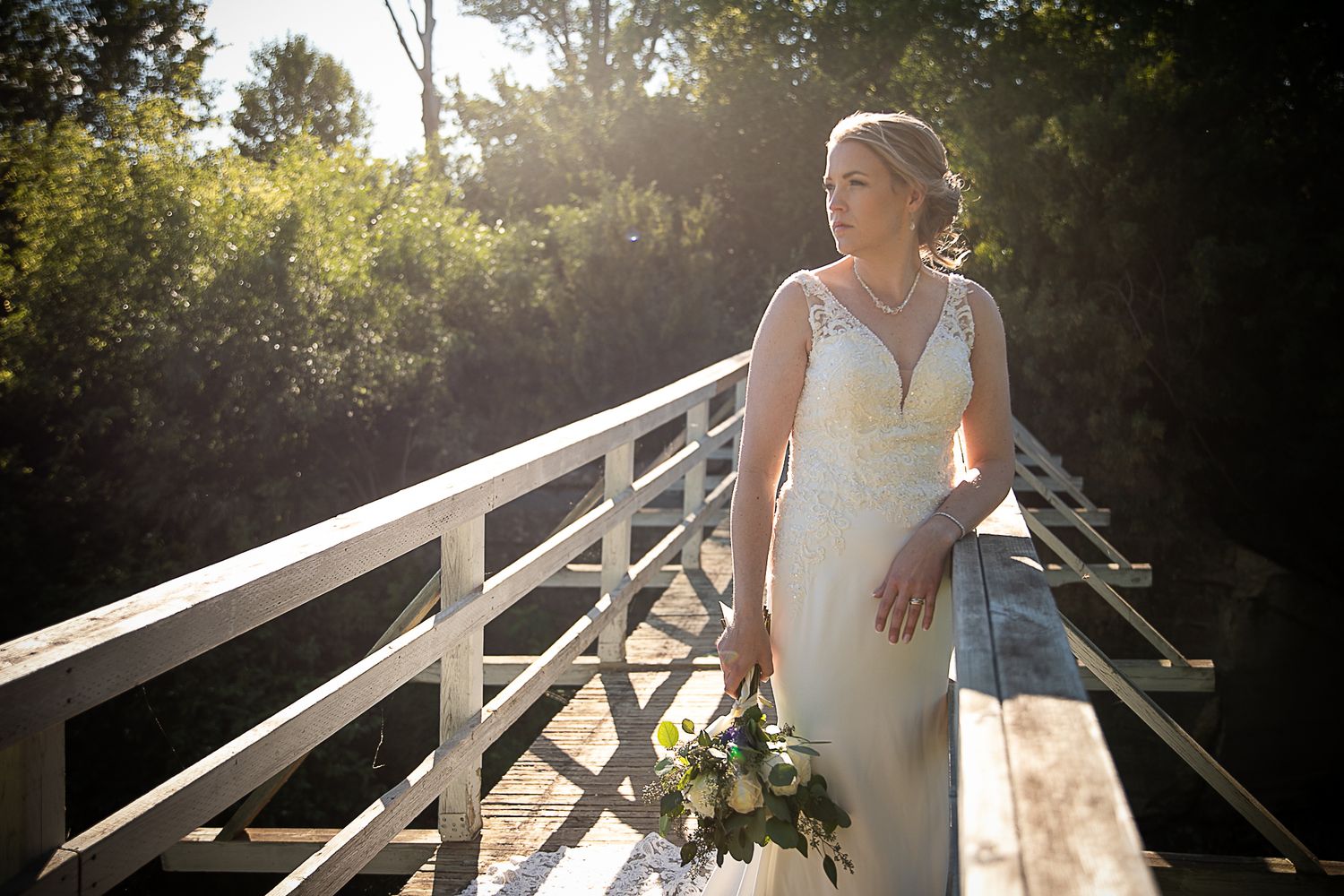 A bride in a fitted white dress stands on a wooden bridge holding a bouquet in golden sunlight.