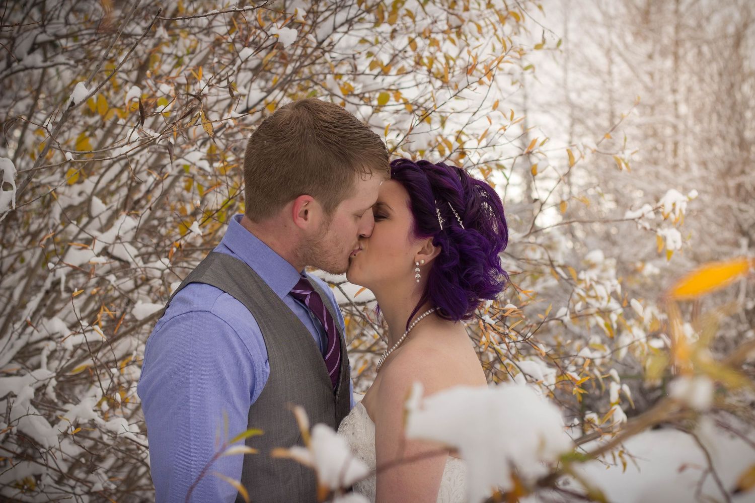 Newlyweds share intimate moments among snow-dusted autumn branches.