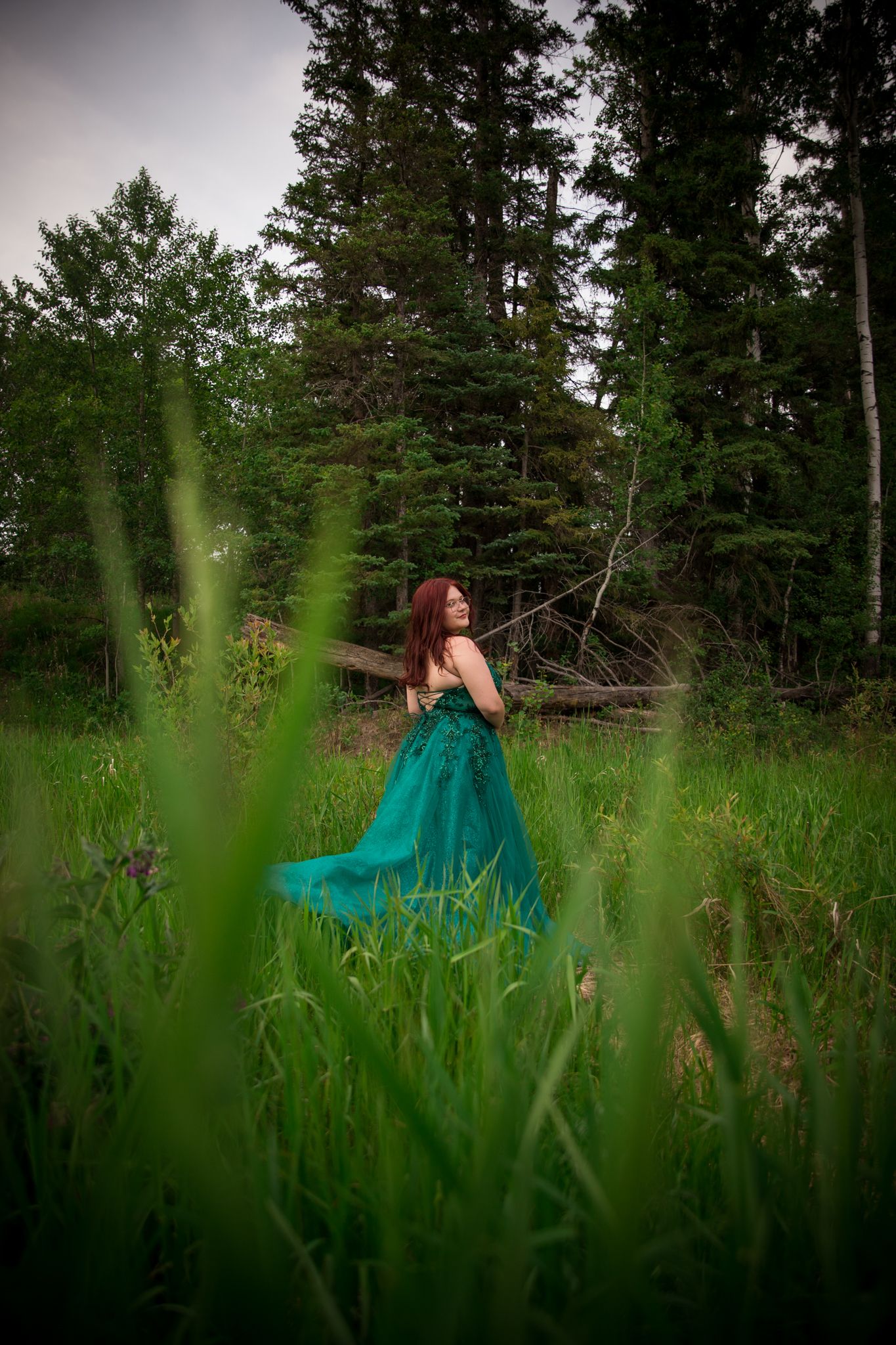 Emerald gown contrasts with tall green grass and pine trees in natural outdoor setting.