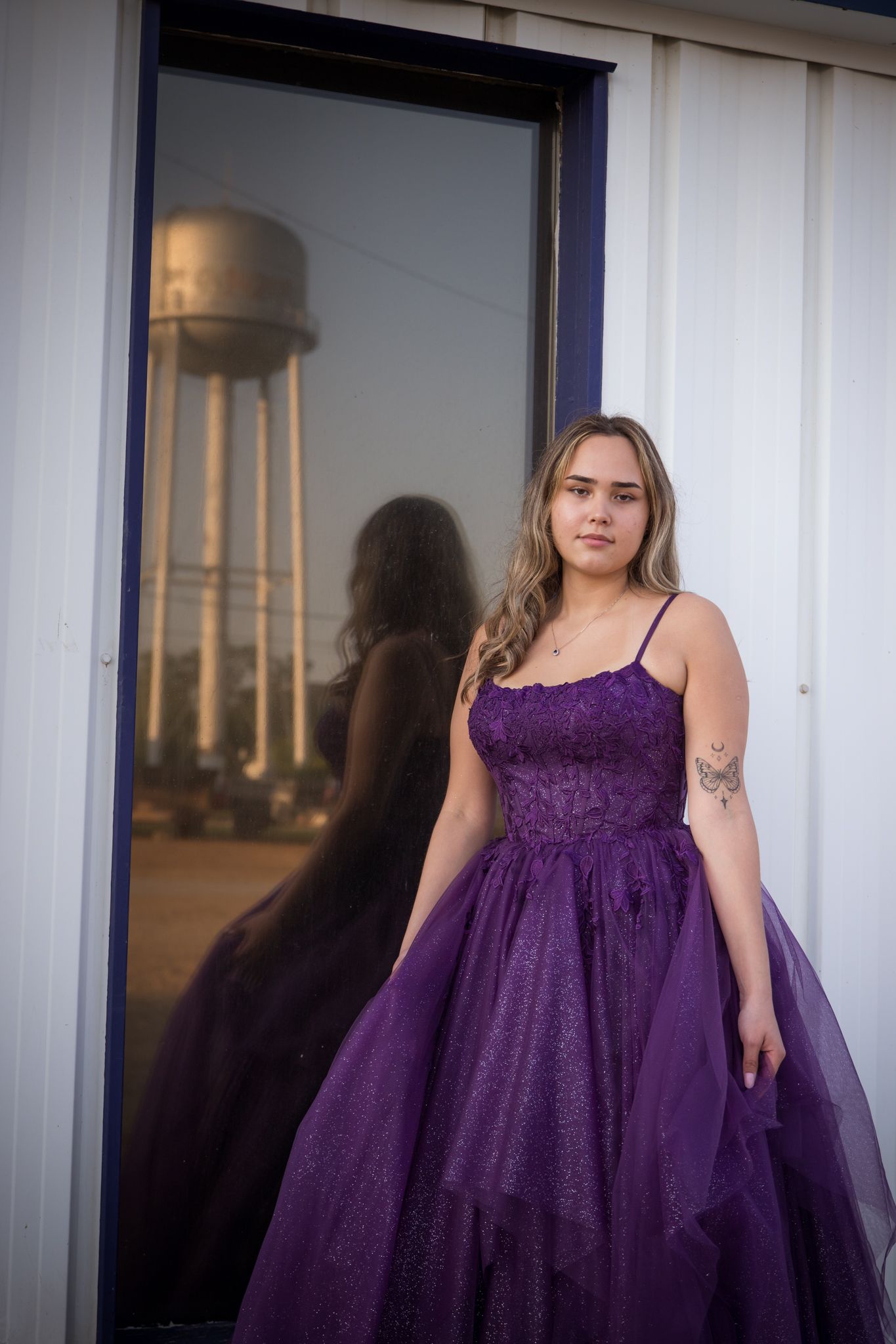 A person in a purple tulle gown poses against an industrial backdrop with a water tower visible.