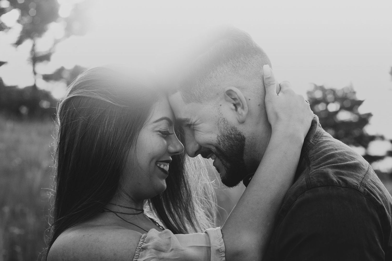 Black and white photo of a laughing couple touching foreheads romantically outdoors in a sunlit field.