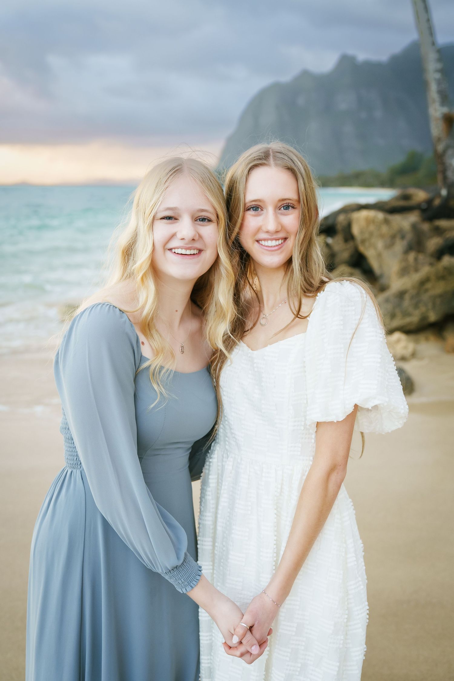 Two sisters in dresses hugging and smiling for the camera at sunrise. Ka’a’awa Beach, Oahu, with mountains and ocean in the background